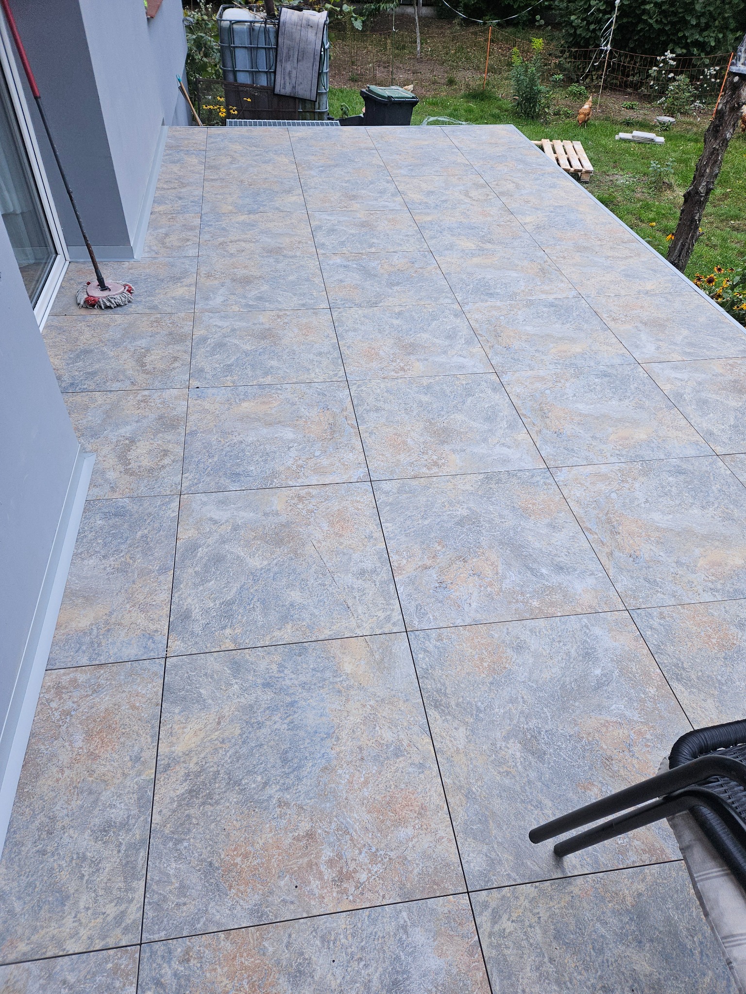 Wyłożony płytkami taras przy domu z mopem w rogu. Szare płytki z beżowymi akcentami. Widok na ogród z paletą i kurami. Część krzeseł w prawym dolnym rogu.