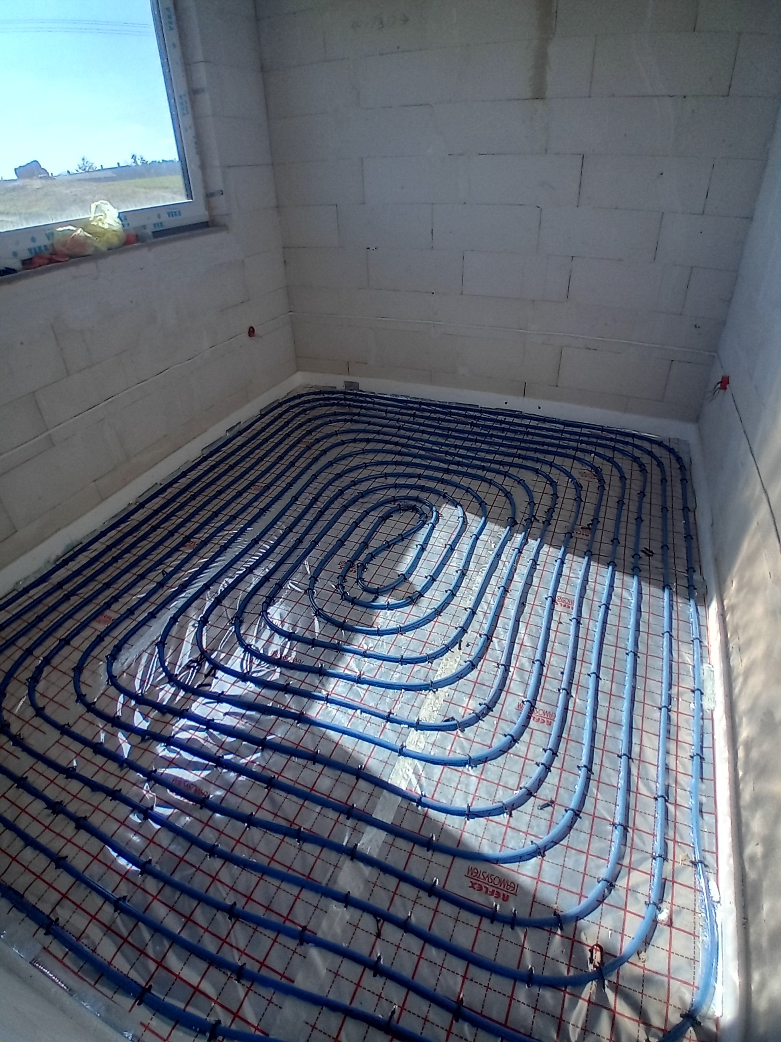 Niebieskie rury ogrzewania podłogowego ułożone spiralnie na folii z siatką w surowym pomieszczeniu z oknem.