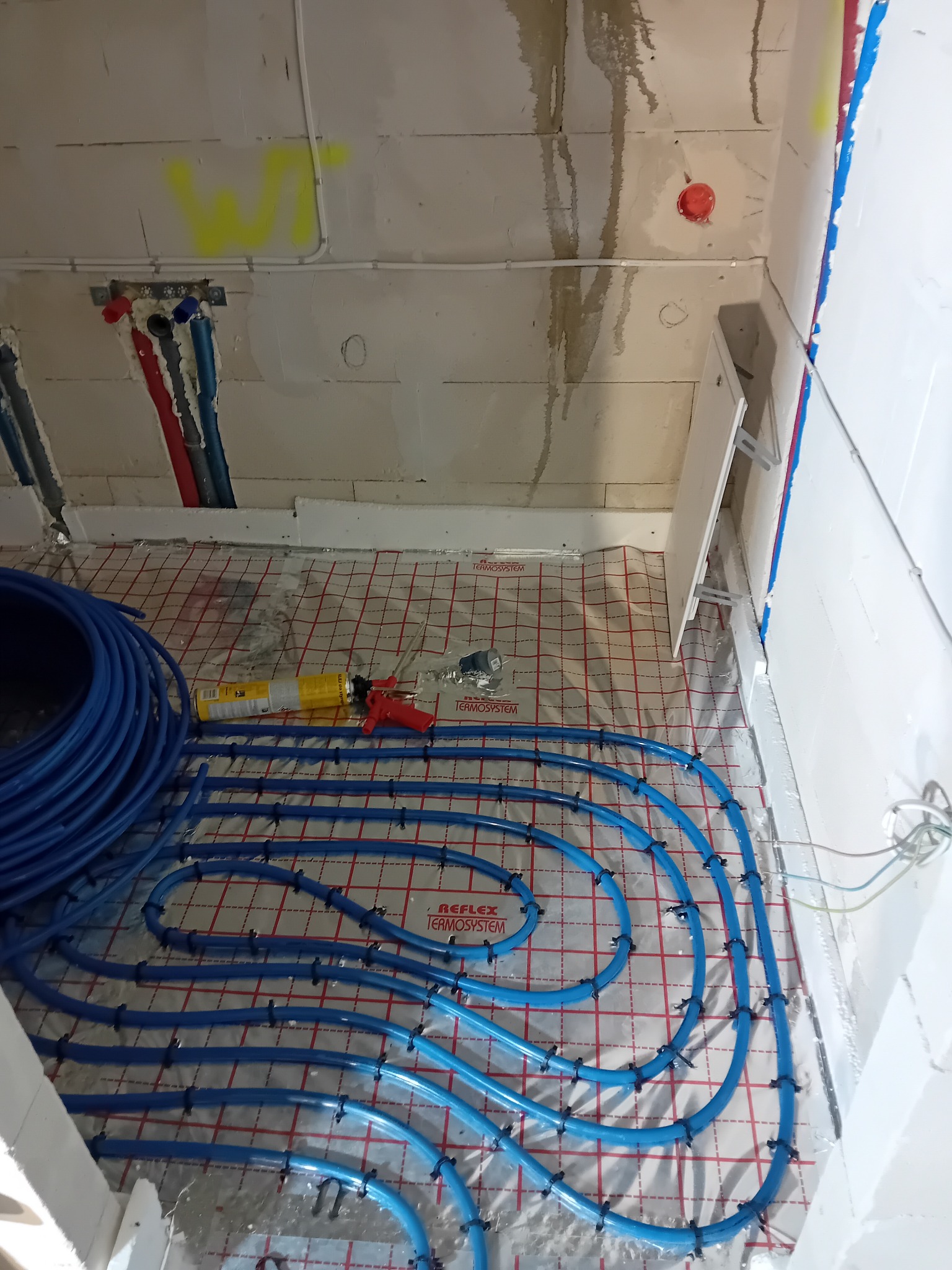 Niedokończona instalacja ogrzewania podłogowego: niebieskie rury ułożone na folii refleksyjnej z widoczną pianką montażową i narzędziami, surowe ściany w tle.