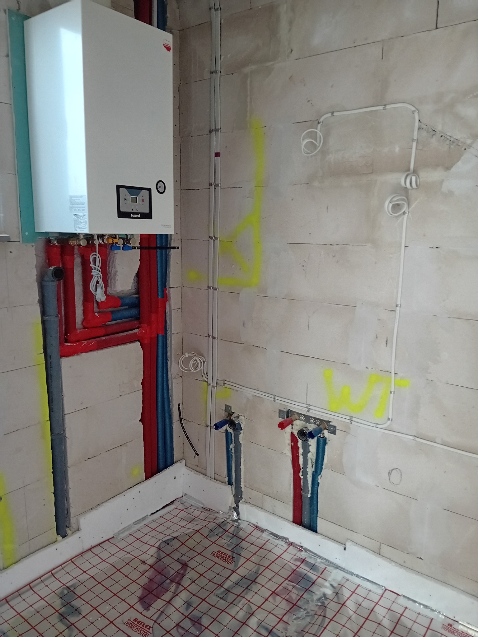 Wnętrze w trakcie instalacji: piec gazowy Termet na ścianie z bloczków, piony instalacyjne w kolorach czerwonym i niebieskim, rury ogrzewania podłogowego na podłodze, widoczne przewody elektryczne...