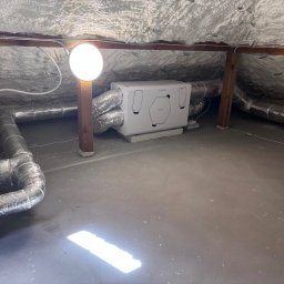 Paweł Czarnecki, Czarnecki Wentylacje - Biała centrala rekuperacyjna z podłączonymi przewodami wentylacyjnymi na poddaszu ocieplonym pianą, oświetlona lampą. Widoczne elementy instalacji i konstrukcji dachu.