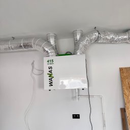 Paweł Czarnecki, Czarnecki Wentylacje - Biała jednostka rekuperacji WANAS 415 HI-TECH z podłączonymi srebrnymi przewodami wentylacyjnymi na białej ścianie. Widoczne rury odpływowe i przewody elektryczne.