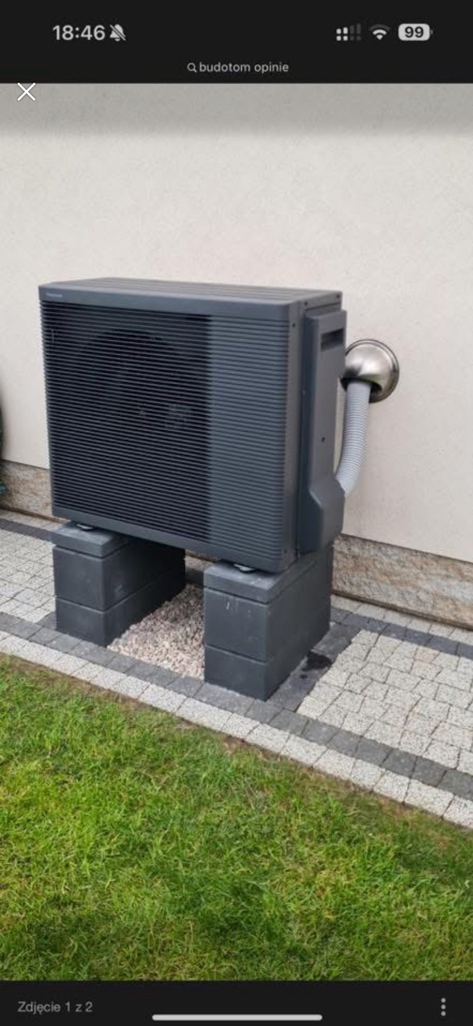 Szara pompa ciepła Panasonic Aquarea zamontowana na betonowych blokach przy ścianie budynku, z widocznym fragmentem trawnika i kostki brukowej.