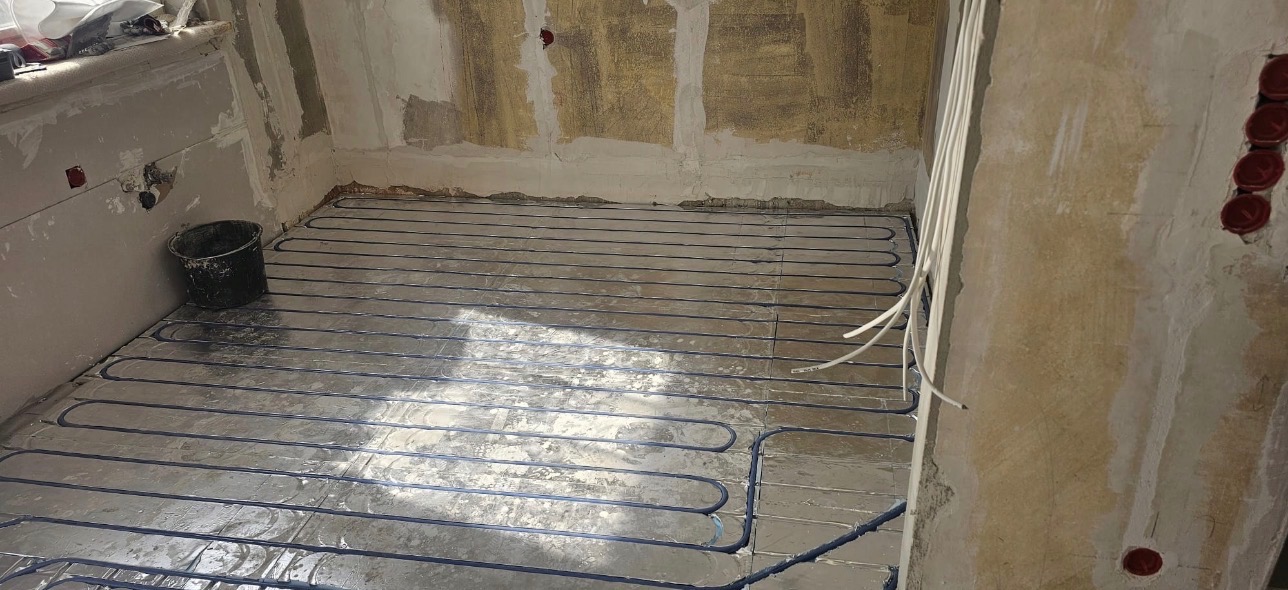 Instalacja ogrzewania podłogowego w trakcie prac wykończeniowych. Widoczne rury grzewcze ułożone na folii izolacyjnej, ściany w stanie surowym, przygotowane do tynkowania.