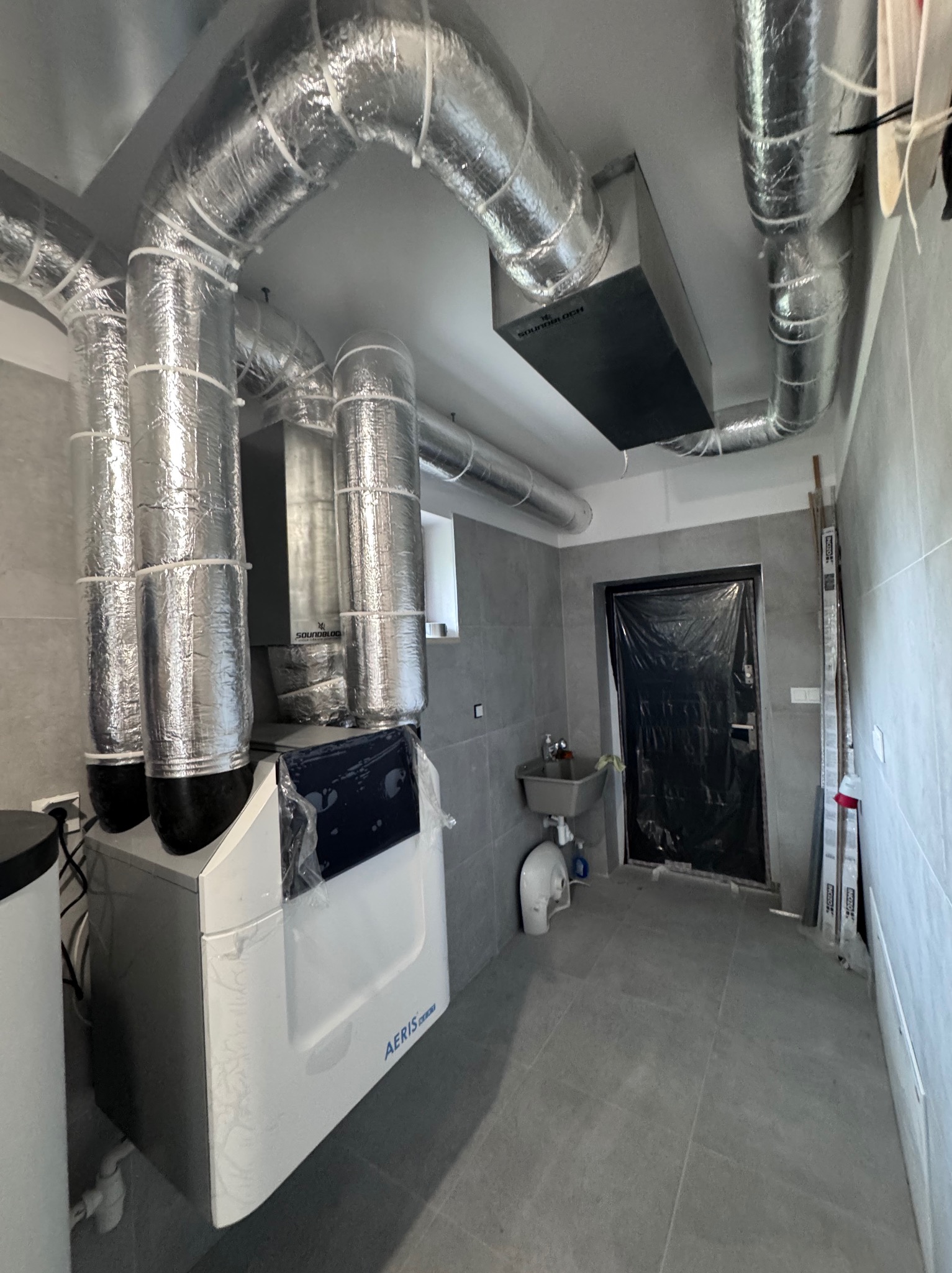 Zmontowana instalacja rekuperacji z rurami w srebrnej izolacji, widoczny rekuperator AERIS, szare płytki na ścianach i podłodze, pomieszczenie w trakcie prac wykończeniowych.