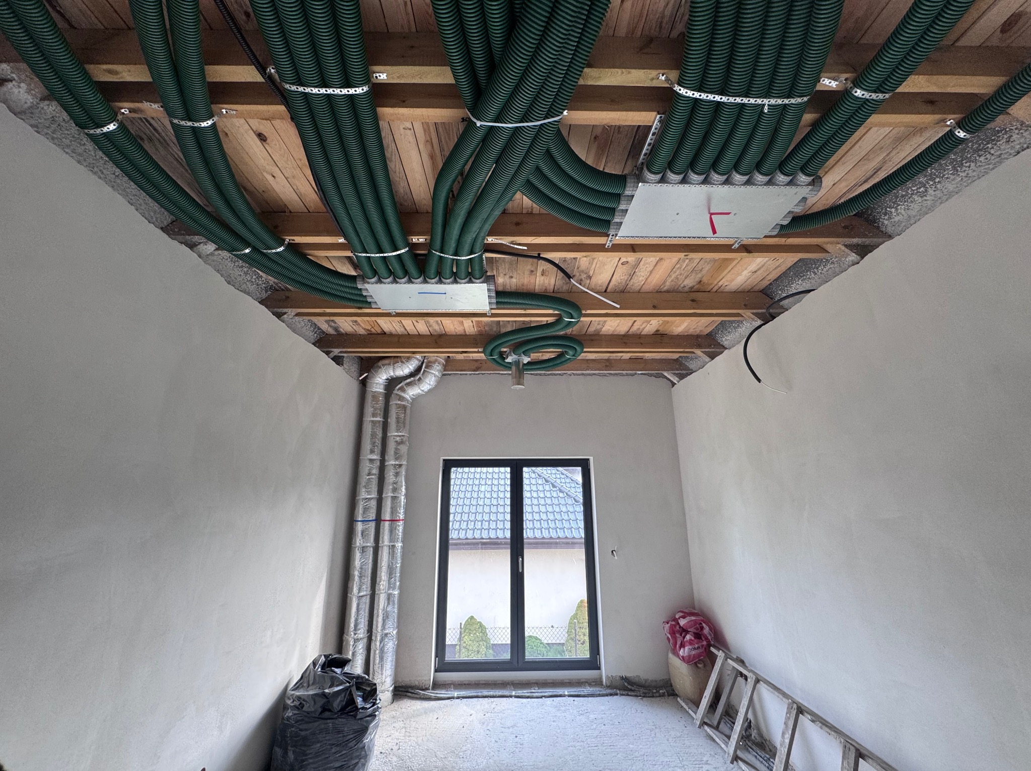 Zielone rury wentylacyjne zamontowane pod drewnianym stropem w surowym wnętrzu. Widoczne puszki rozdzielcze i fragment instalacji przy oknie. Prace instalacyjne w trakcie.