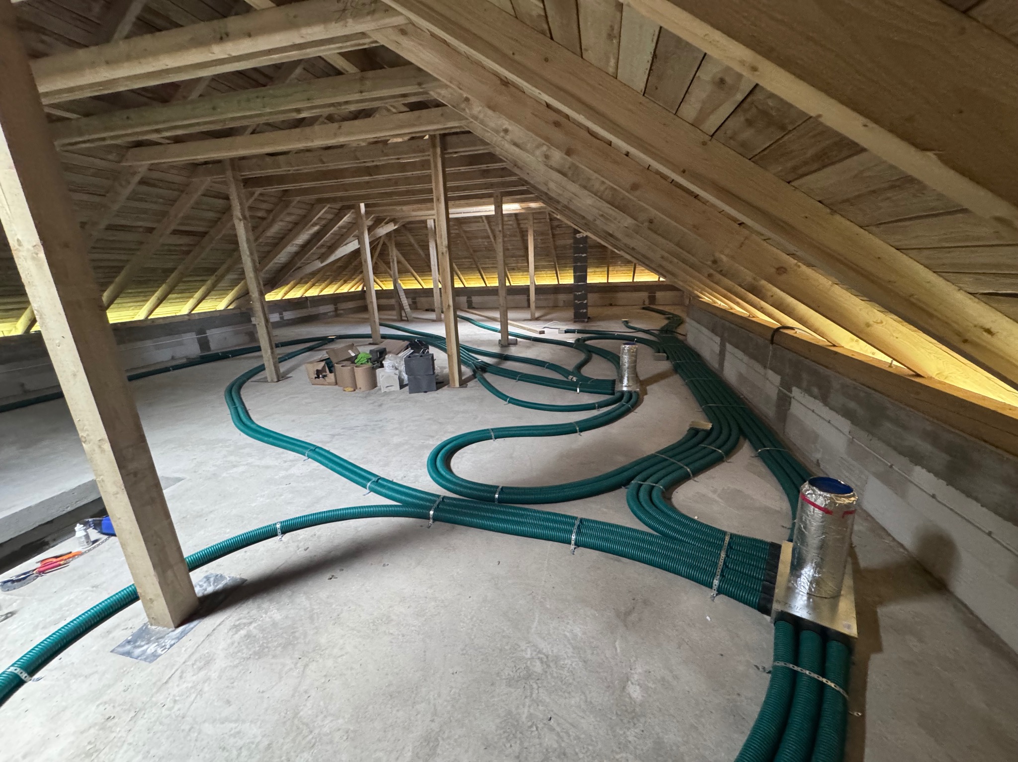 Zielone rury wentylacyjne ułożone w surowym wnętrzu poddasza z drewnianą konstrukcją dachu. Widoczne narzędzia i materiały instalacyjne.