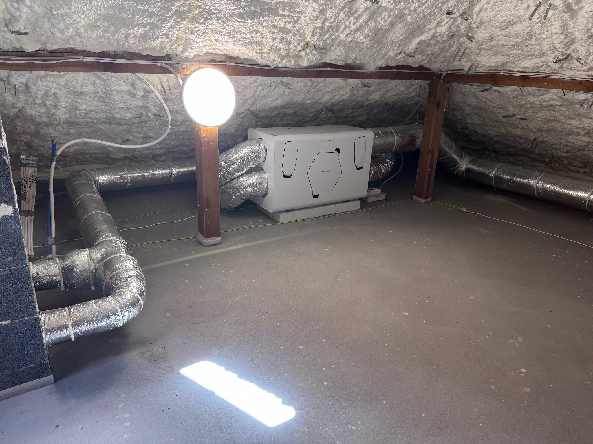 Biała centrala rekuperacyjna z podłączonymi przewodami wentylacyjnymi na poddaszu ocieplonym pianą, oświetlona lampą. Widoczne elementy instalacji i konstrukcji dachu.