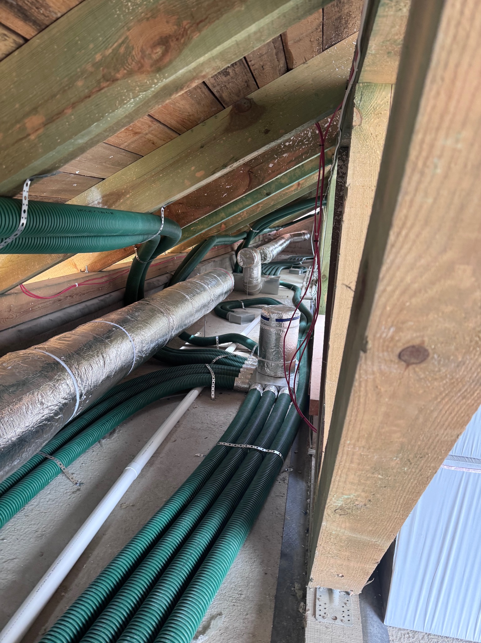 Zielone rury wentylacyjne i srebrny przewód izolowany, zamontowane pod drewnianą konstrukcją dachu. Widoczne elementy mocujące i kable elektryczne.