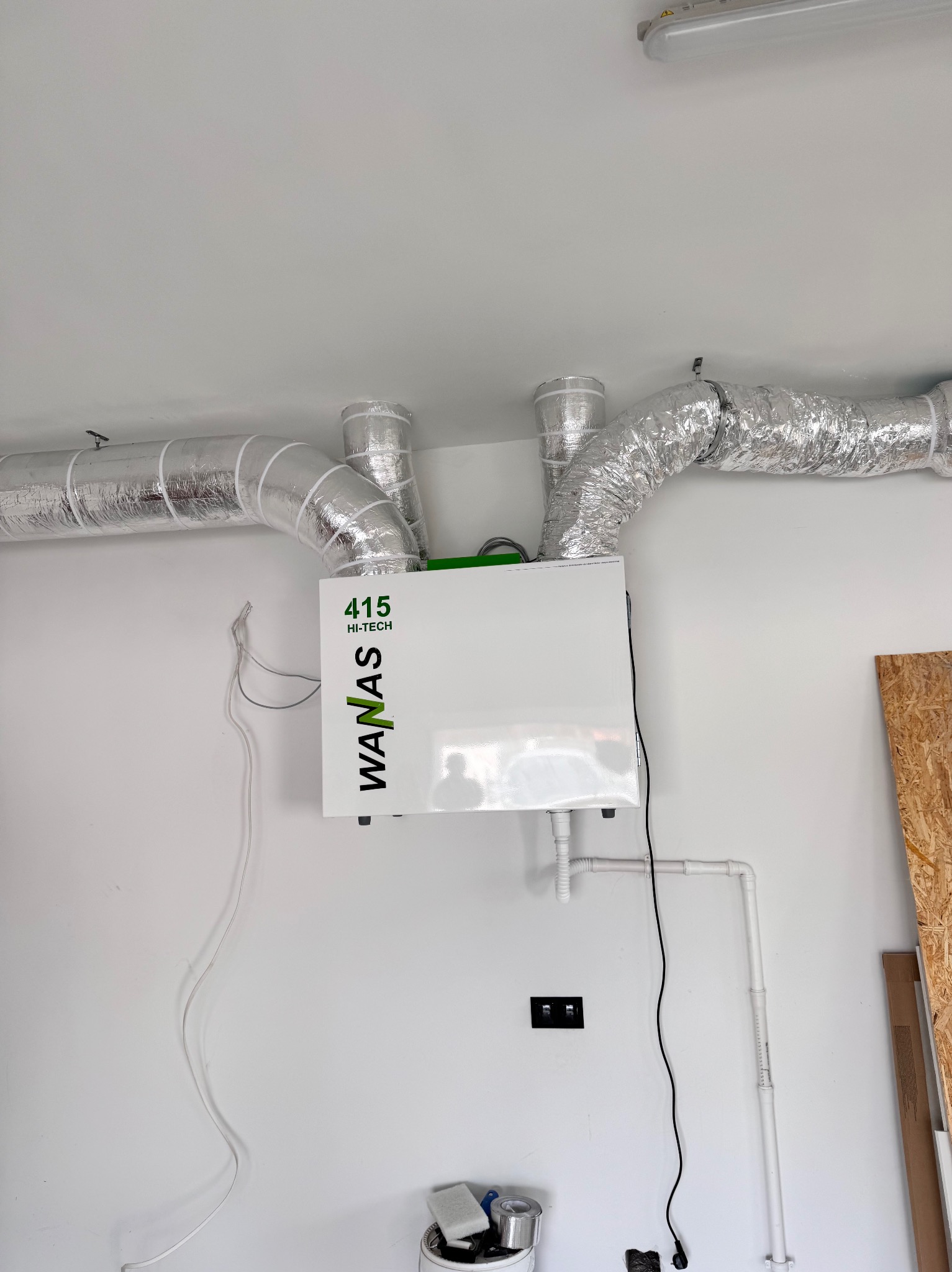 Biała jednostka rekuperacji WANAS 415 HI-TECH z podłączonymi srebrnymi przewodami wentylacyjnymi na białej ścianie. Widoczne rury odpływowe i przewody elektryczne.