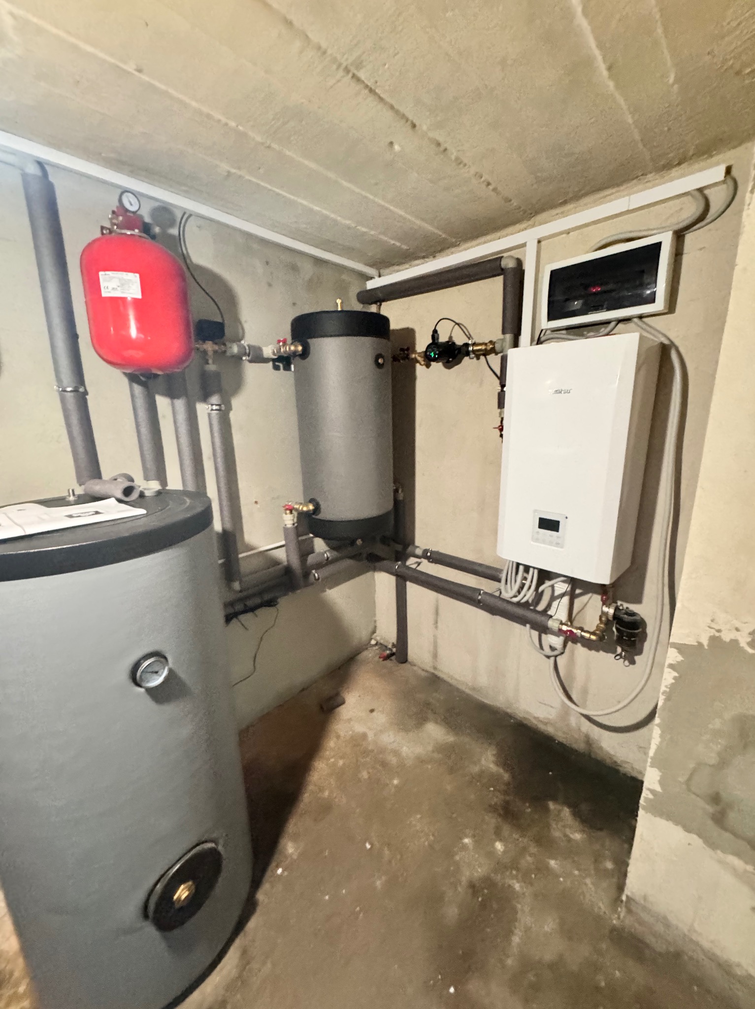 Zestaw urządzeń grzewczych w kotłowni: zbiorniki akumulacyjne, ekspansomat, piec gazowy kondensacyjny i sterownik. Szare rury izolowane. Surowe ściany i podłoga.