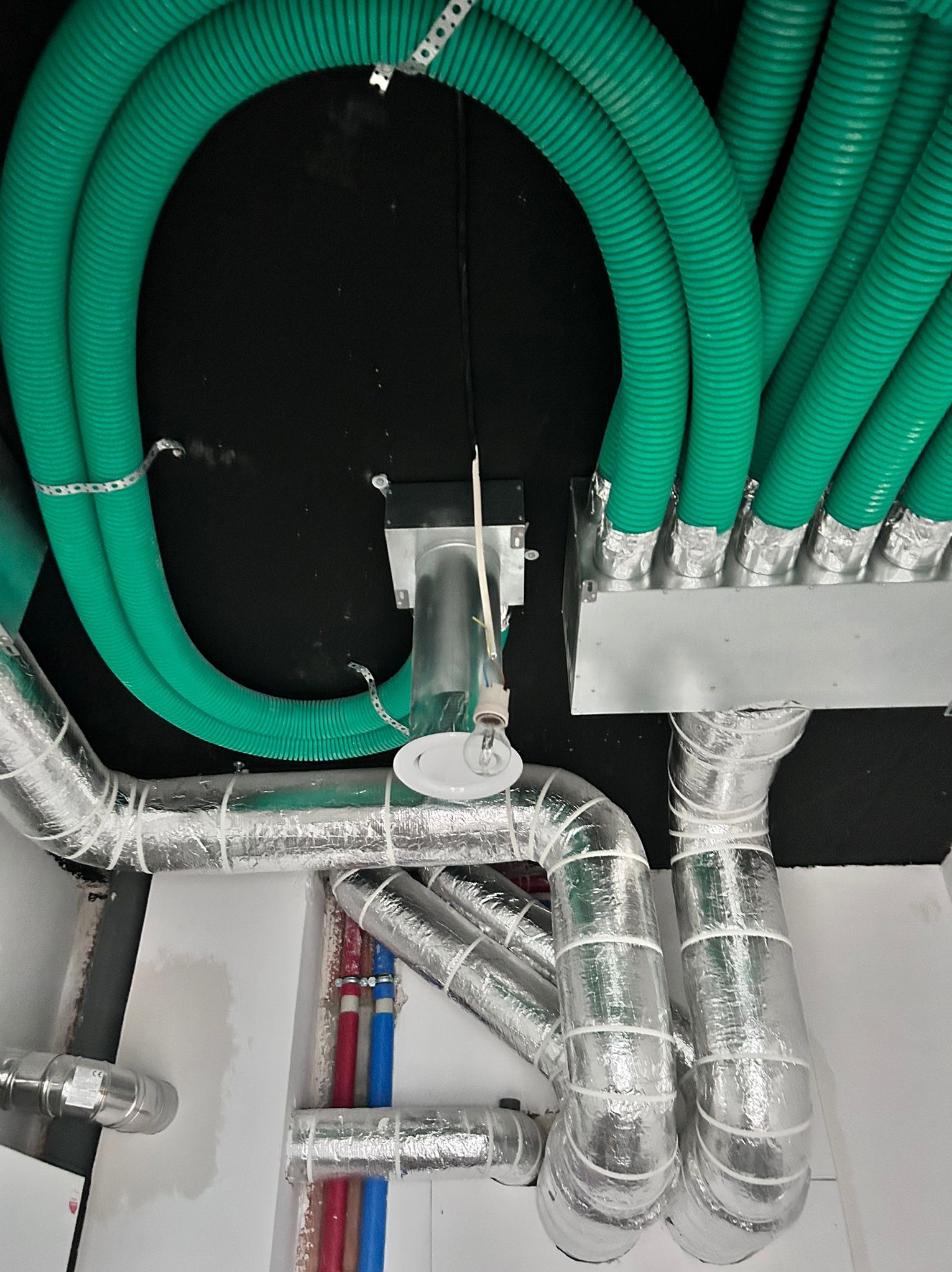 Zielone i srebrne rury wentylacyjne pod sufitem, widoczne elementy instalacji, w tym rury w izolacji i skrzynka rozdzielcza. Ujęcie z dołu, instalacja w trakcie montażu.