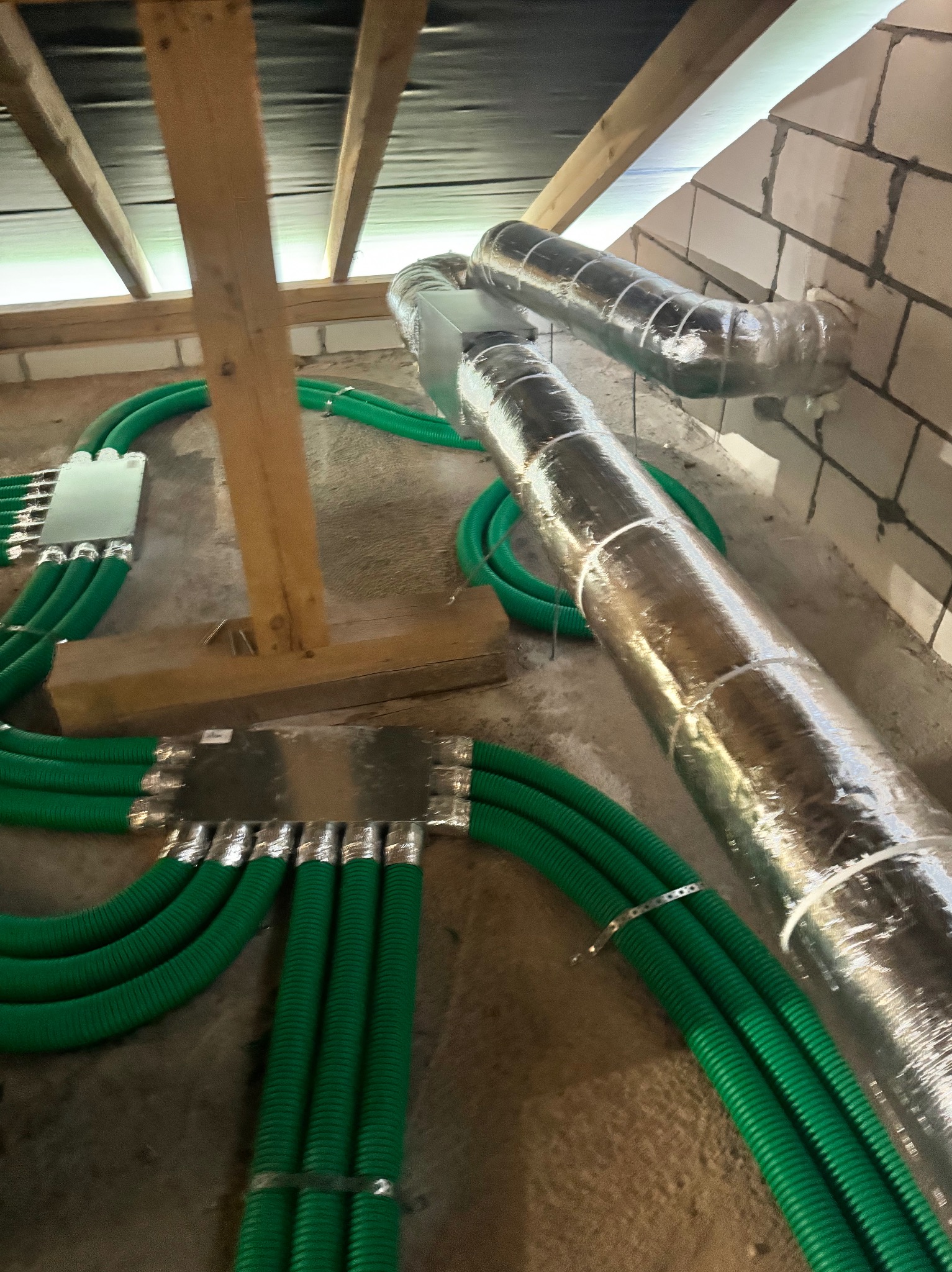 Zielone rury wentylacyjne i srebrne przewody izolowane, zamontowane pod stropem w surowym stanie, widoczne elementy konstrukcji drewnianej i ściany z bloczków.