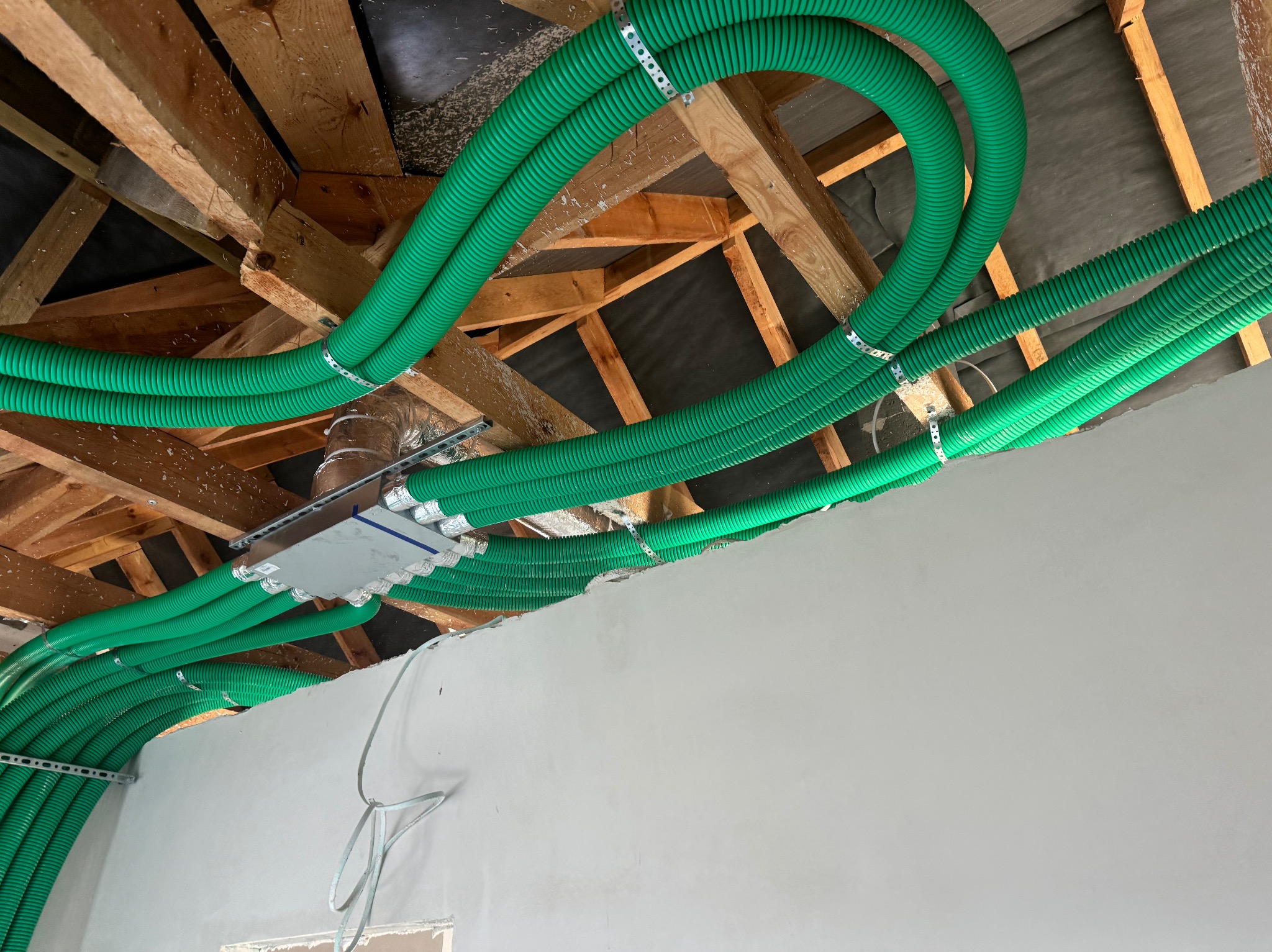 Zielone, karbowane rury wentylacyjne zamontowane pod drewnianą konstrukcją dachu, przymocowane metalowymi obejmami. Widoczny fragment ściany i skrzynka rozdzielcza.