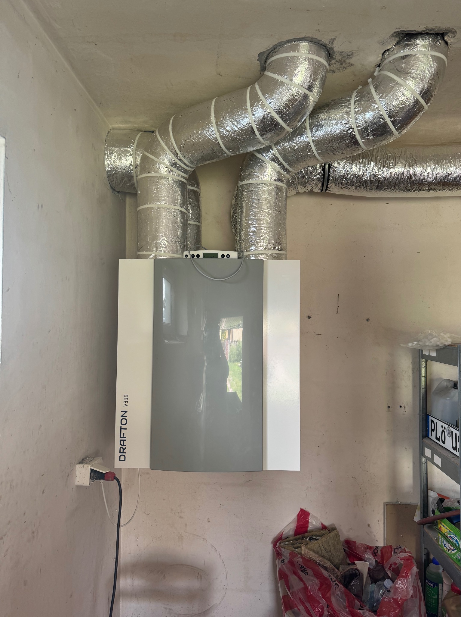 Szara centrala wentylacyjna Drafton V300 z podłączonymi, zaizolowanymi przewodami wentylacyjnymi w surowym pomieszczeniu. Widoczny fragment regału i gniazdko elektryczne.