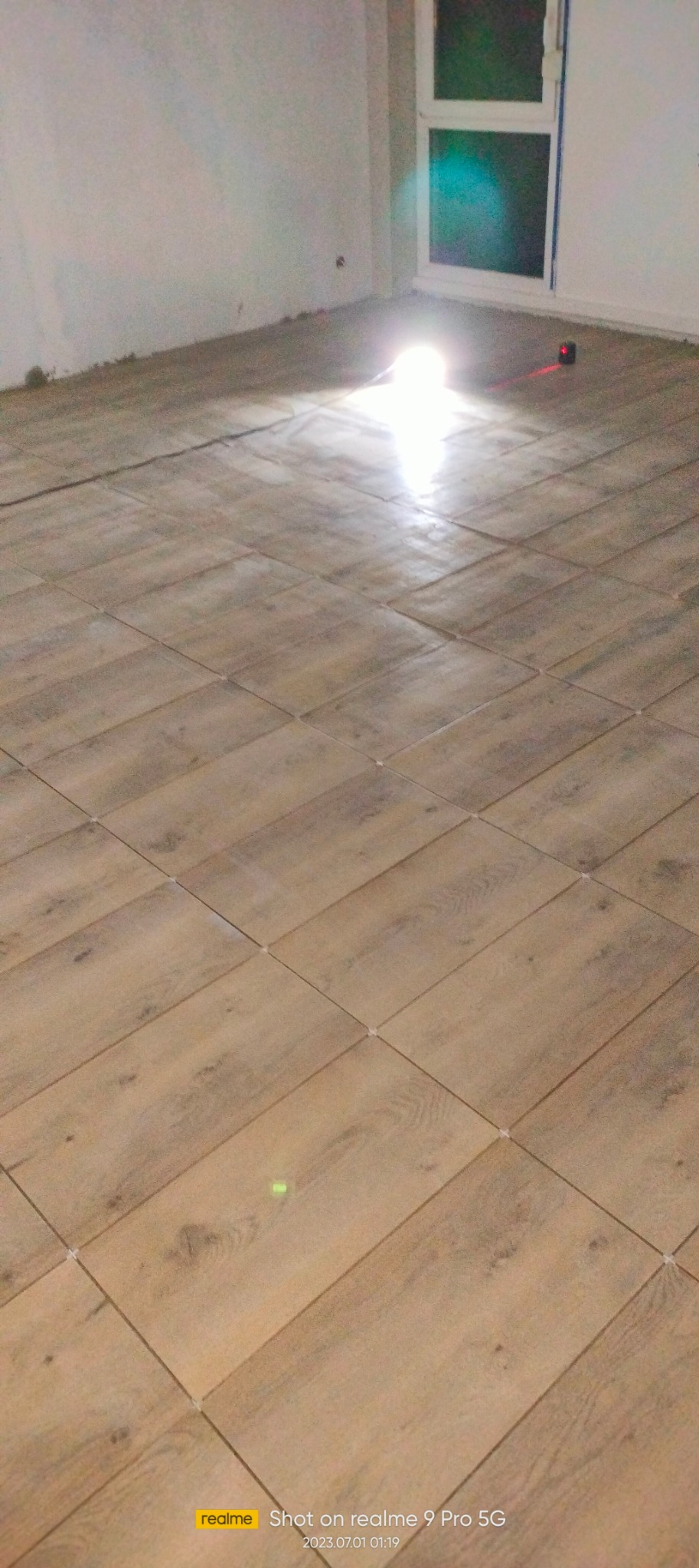 Ułożona podłoga z płytek imitujących drewno, z krzyżykami dystansowymi, oświetlona laserowym niwelatorem. Widok na okno i fragment ściany.