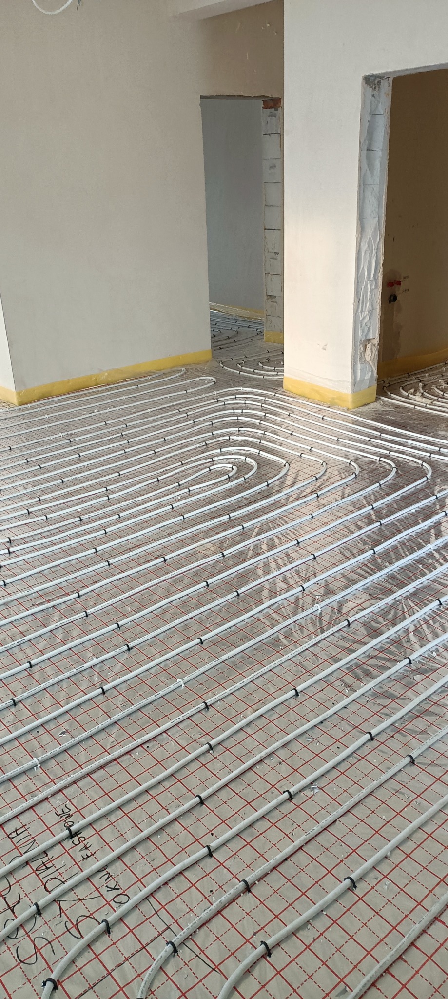 Instalacja ogrzewania podłogowego w trakcie realizacji w Sulechowie. Białe rurki ułożone na czerwonej siatce, przygotowane do zalania posadzką. Widoczne ściany w surowym stanie.