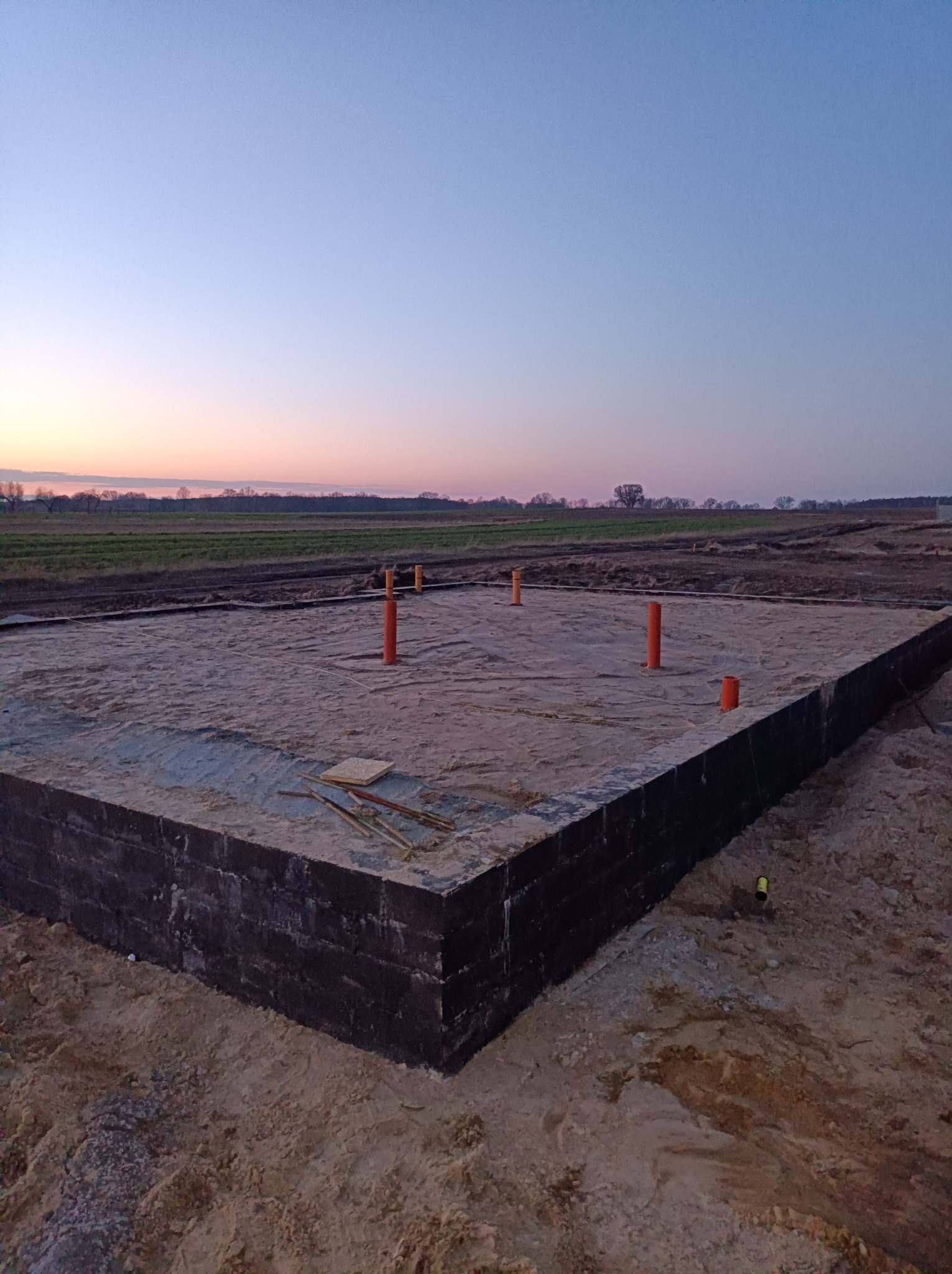 Fundamenty domu jednorodzinnego z wyprowadzonymi pionami instalacji, widoczne ocieplenie fundamentów, tło stanowi pole i zachodzące słońce.