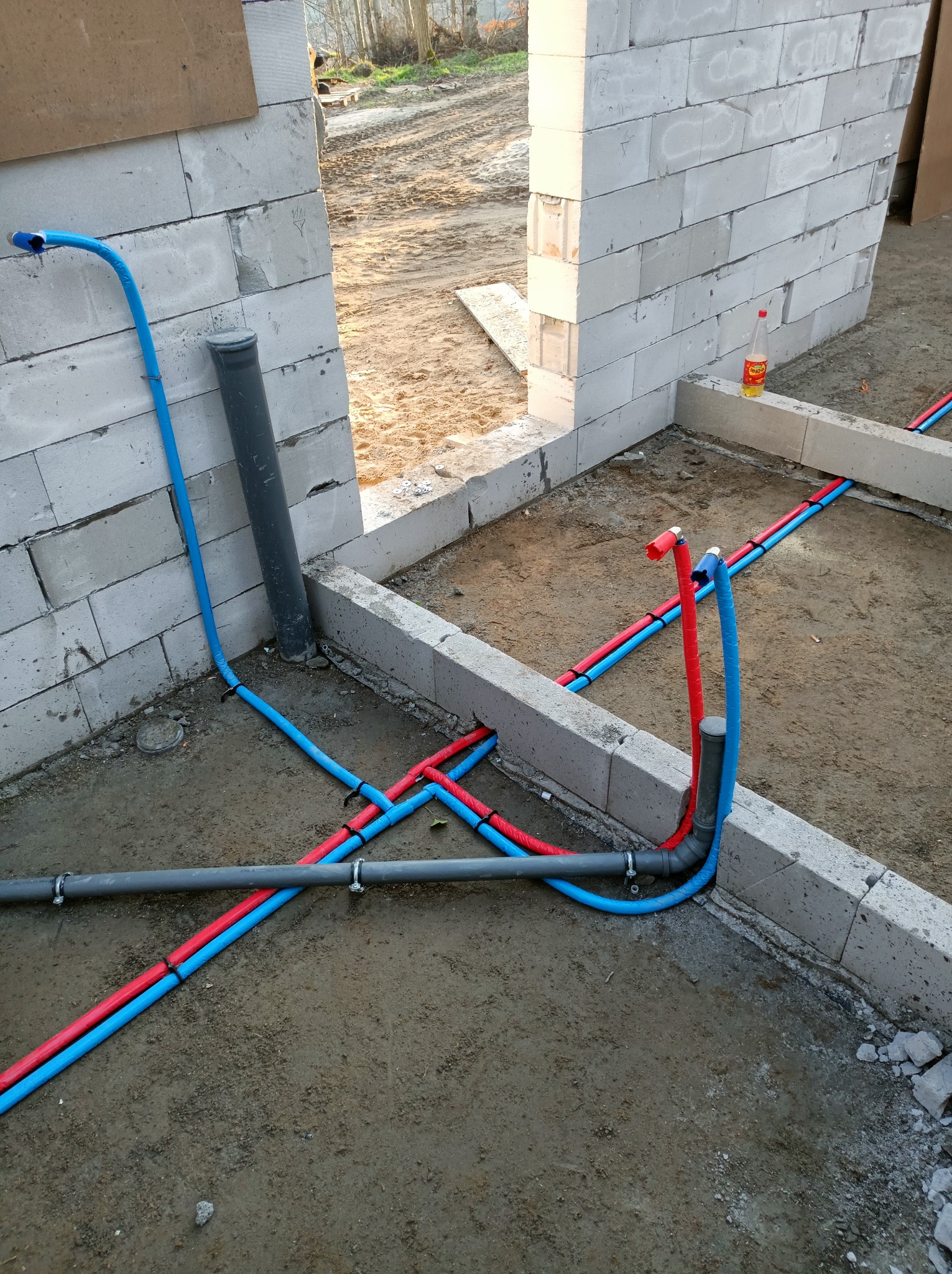 Instalacja wodna PEX: czerwone i niebieskie rury ułożone w surowym stanie, przed wylewką, na tle ściany z betonu komórkowego. Widoczne złączki i mocowania.