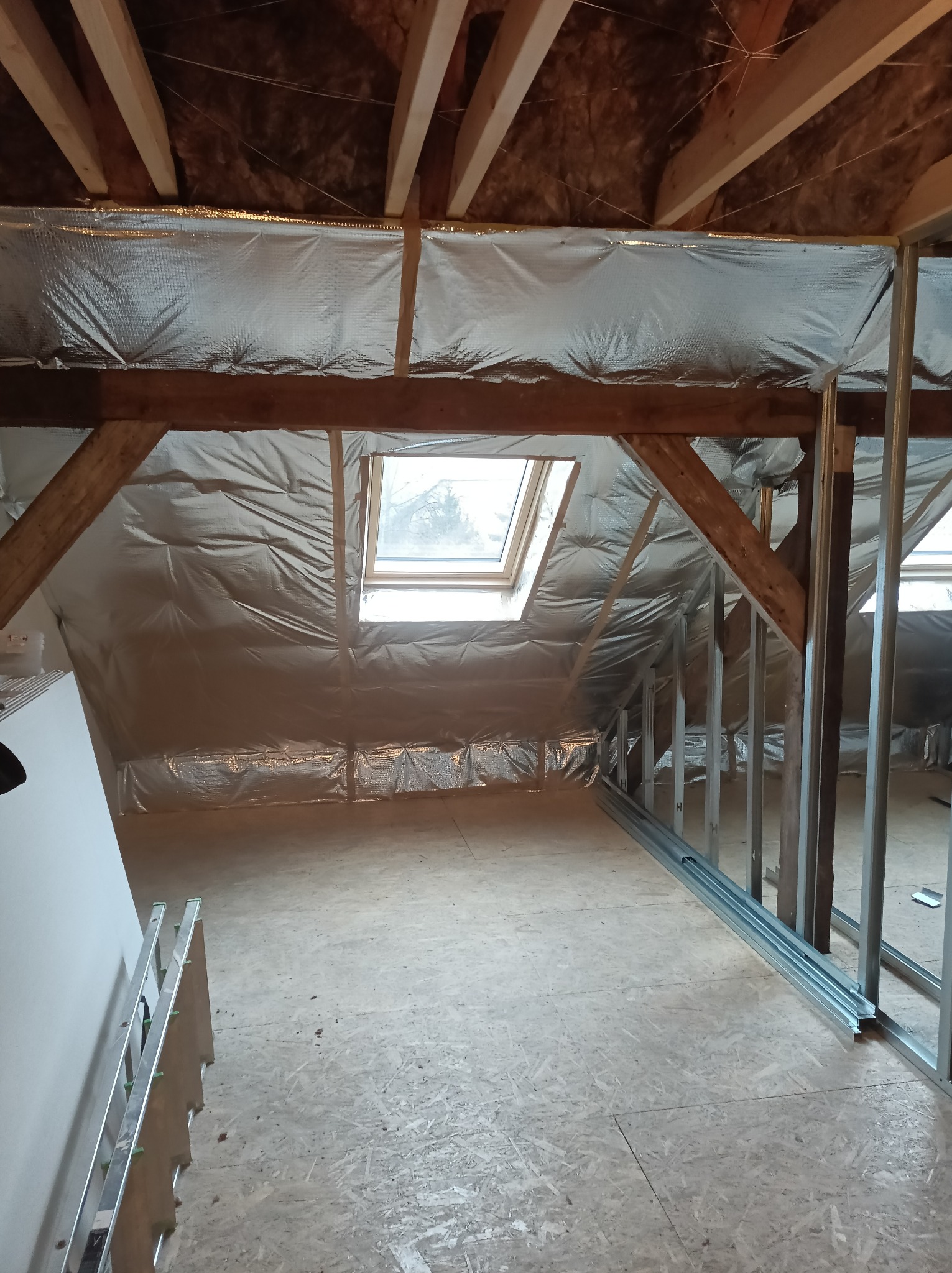 Poddasze z oknem dachowym w trakcie adaptacji: izolacja folią aluminiową, drewniane belki konstrukcyjne, metalowy szkielet ścian działowych, płyty OSB na podłodze.