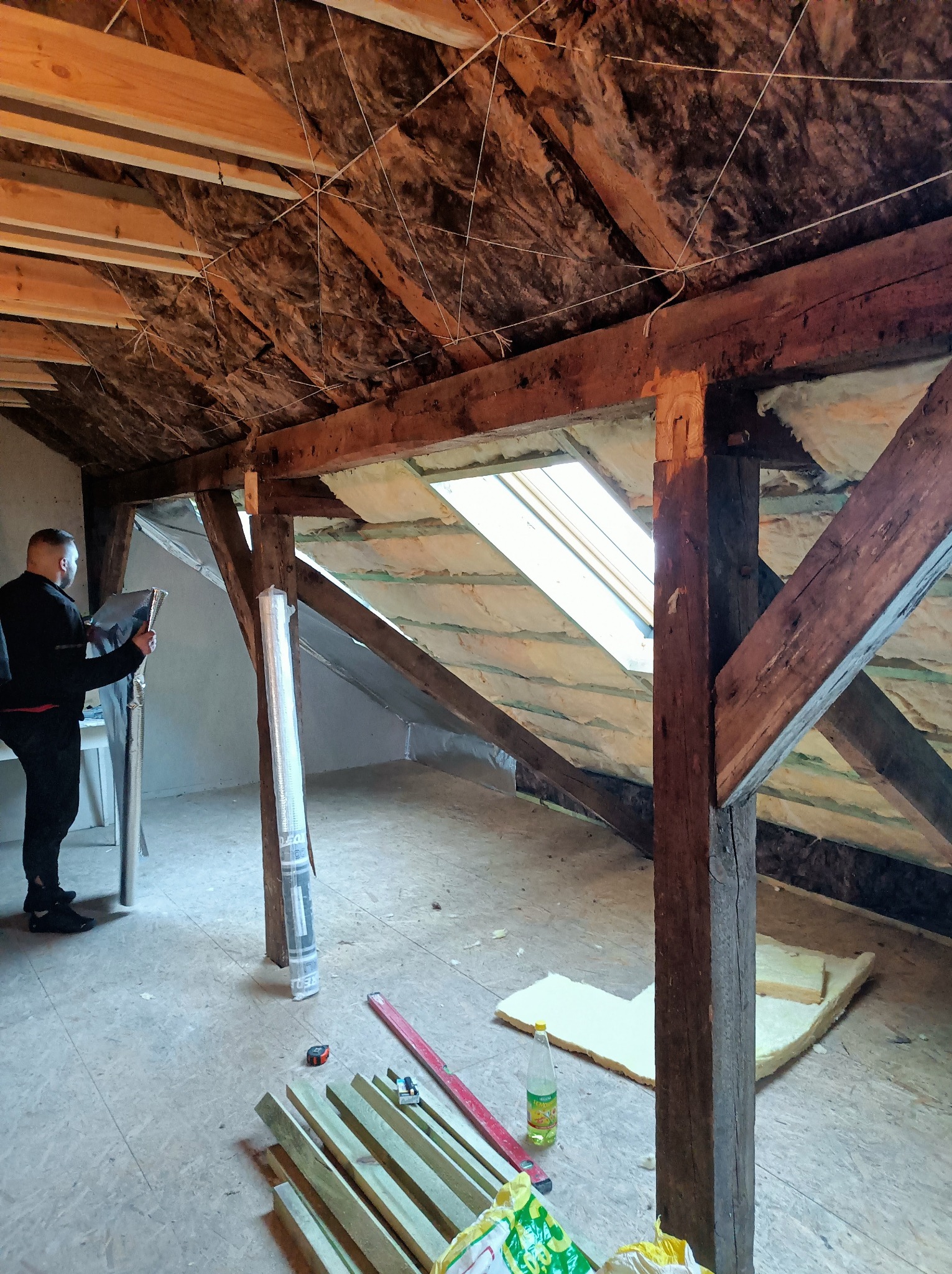 Adaptacja poddasza: ocieplenie dachu wełną mineralną, widoczne drewniane belki konstrukcyjne i okno dachowe. Pracownik w trakcie montażu folii izolacyjnej.