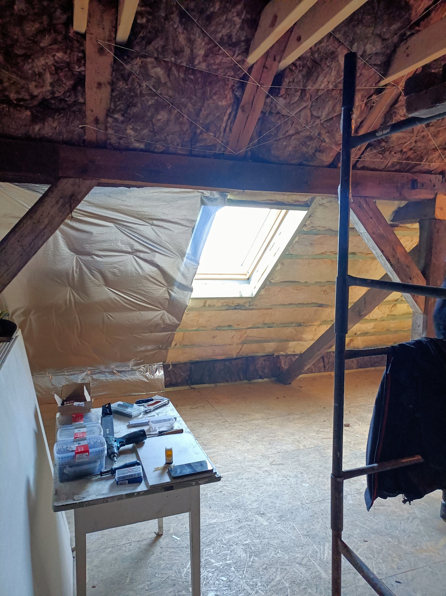 Adaptacja poddasza: widok na izolację folią i wełną mineralną, drewniane belki, okno dachowe, narzędzia na stole i rusztowanie. Prace wykończeniowe.