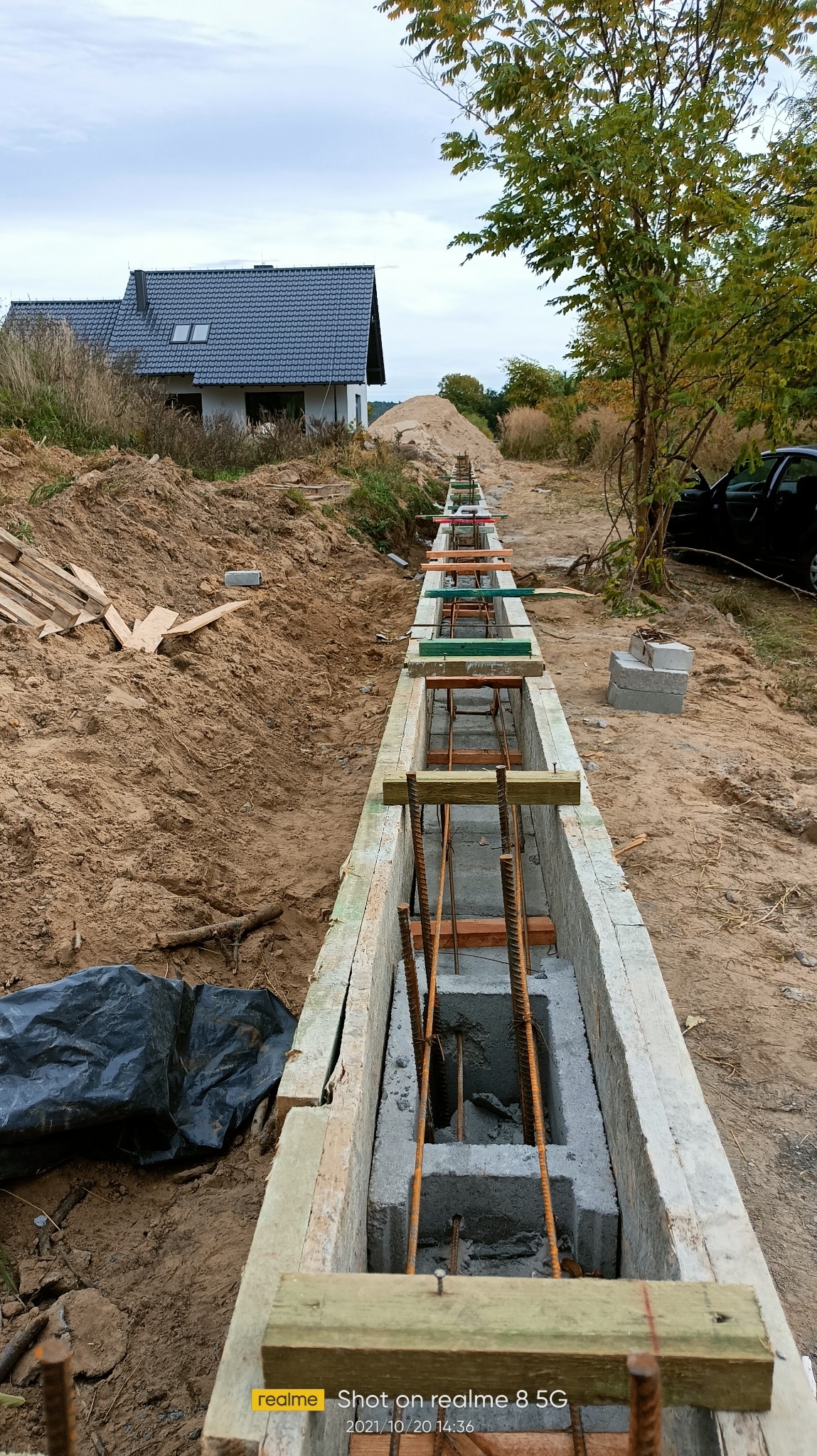 Długi, wąski wykop z betonowymi bloczkami i stalowym zbrojeniem, przygotowany pod fundament domu. Widoczne drewniane szalunki i ziemia wokół.