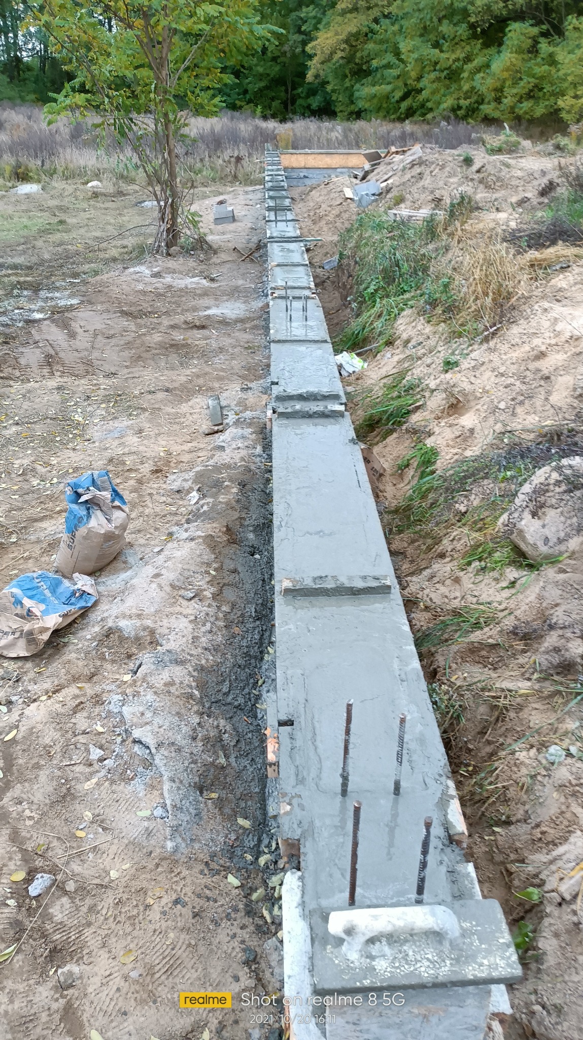 Świeżo wylany fundament z wystającymi prętami zbrojeniowymi, widoczne worki z cementem i drewniane szalunki. Praca w toku, ziemisty teren z roślinnością w tle.