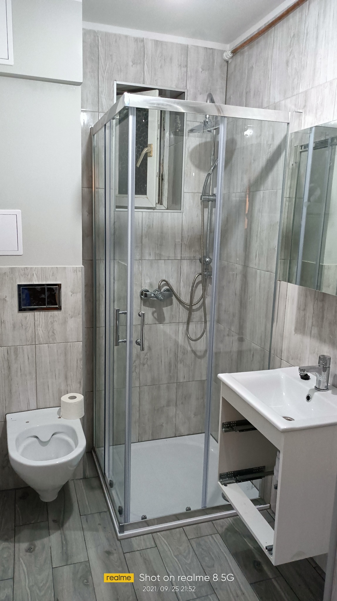 Nowoczesna kabina prysznicowa z armaturą, umywalka z szafką i toaleta w wykończonej łazience. Płytki imitujące drewno na ścianach i podłodze.
