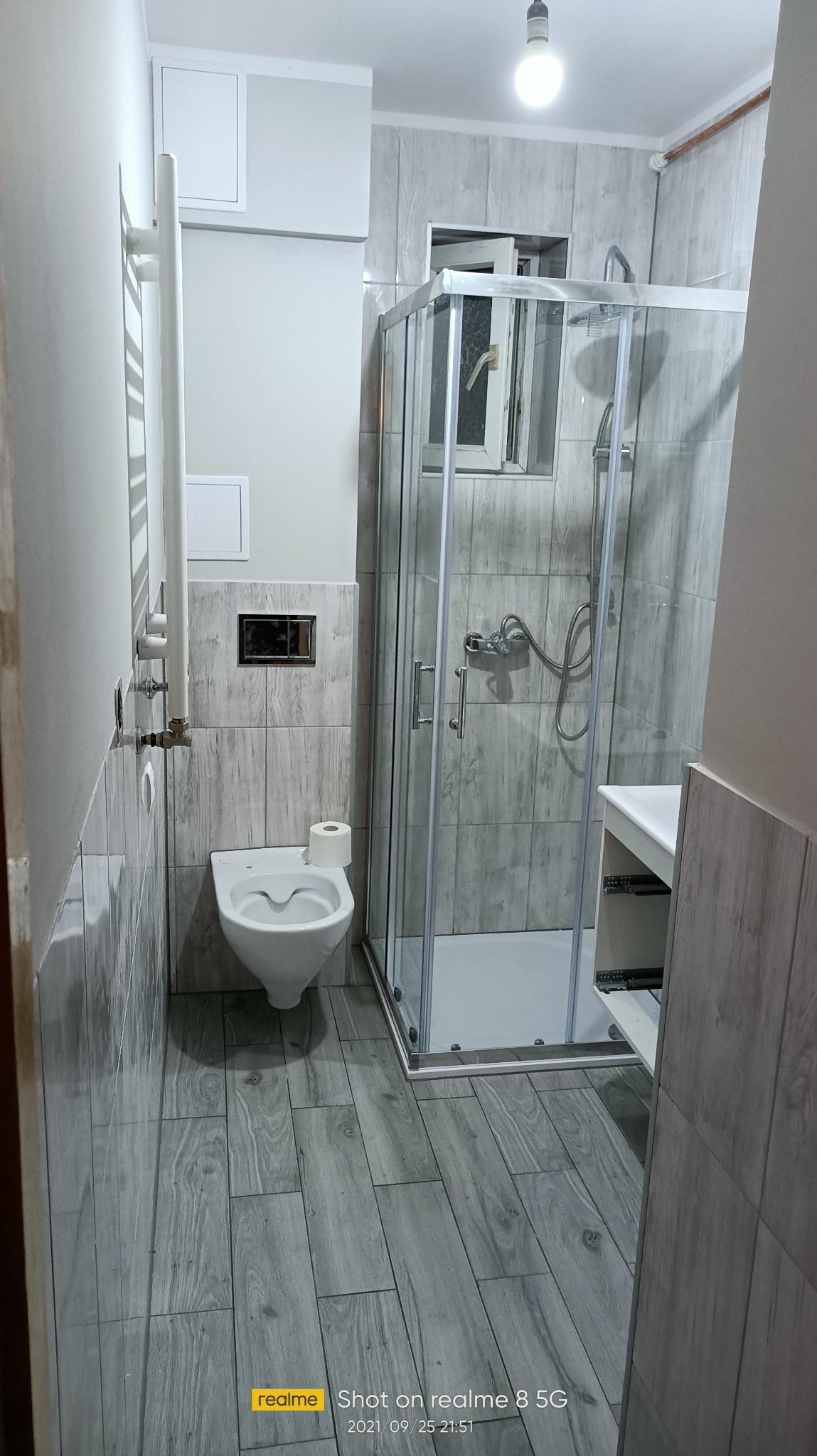 Wykończona łazienka z kabiną prysznicową, toaletą i grzejnikiem drabinkowym. Płytki imitujące drewno na ścianach i podłodze. Nowoczesny design, szara kolorystyka.