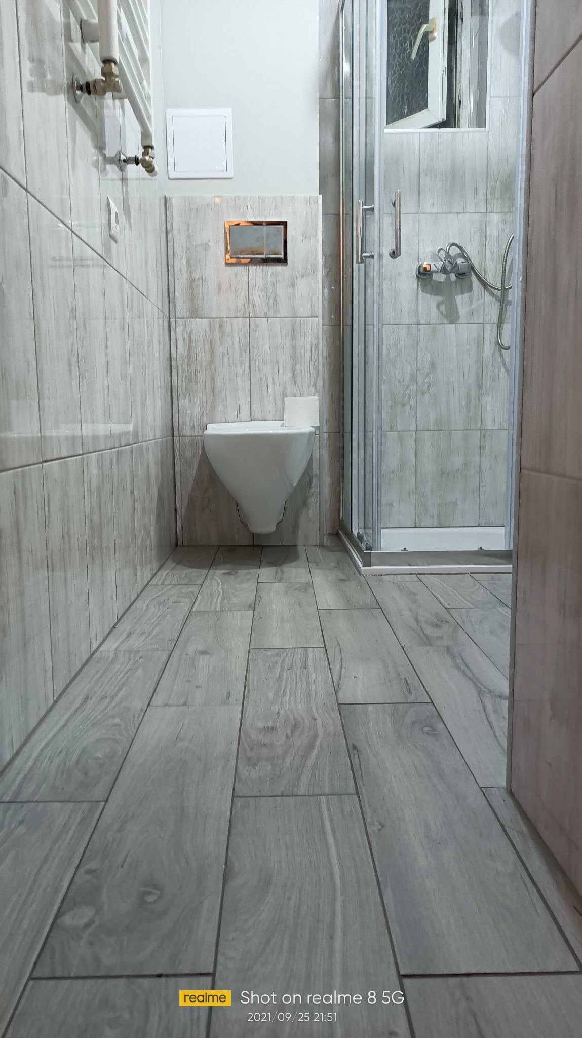 Wykończona łazienka z kabiną prysznicową i podwieszaną toaletą. Płytki na podłodze i ścianach imitują drewno. Widoczny grzejnik drabinkowy i akcesoria łazienkowe.