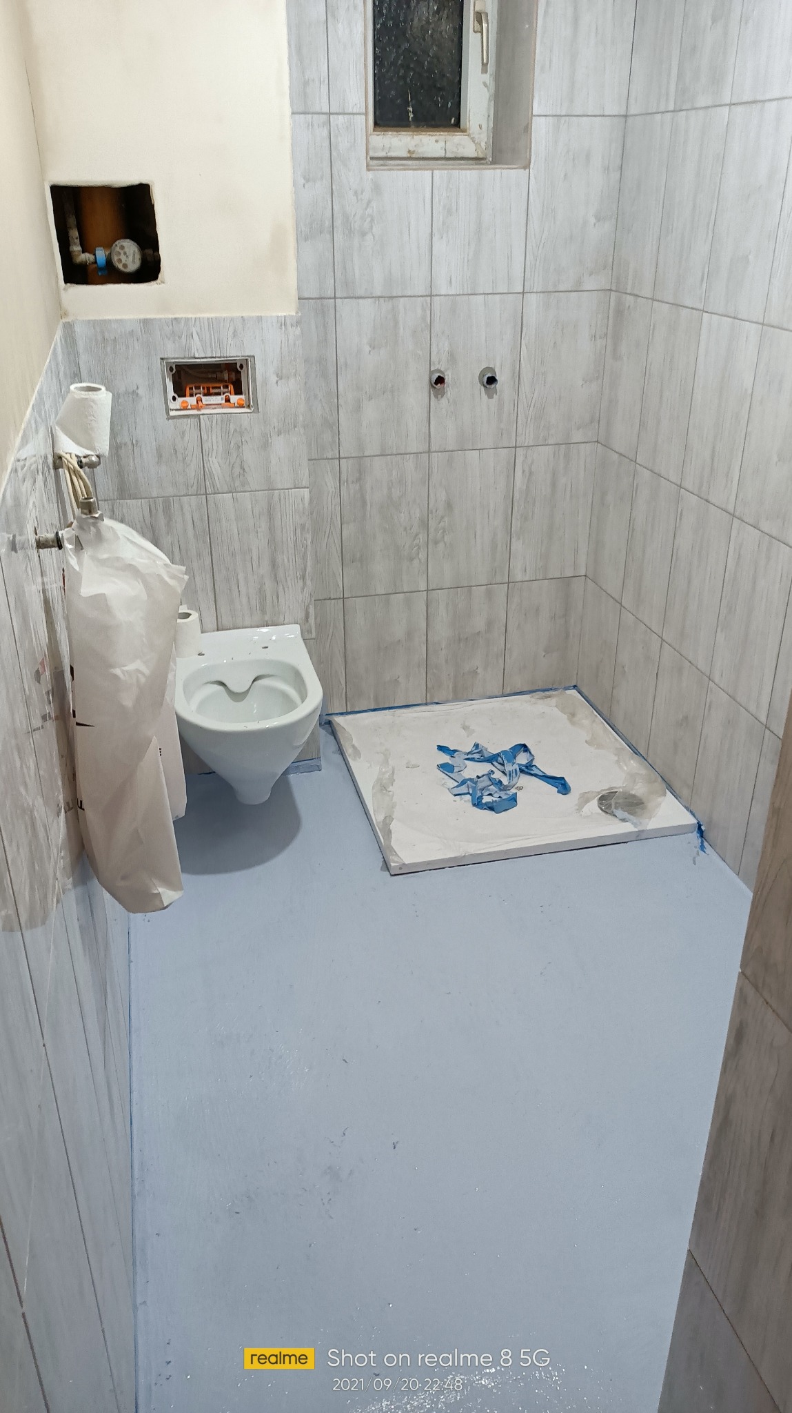 Wyremontowana łazienka z nową, jasną glazurą, pomalowaną podłogą na niebiesko i zamontowaną toaletą. Widoczne przygotowania do montażu kabiny prysznicowej.