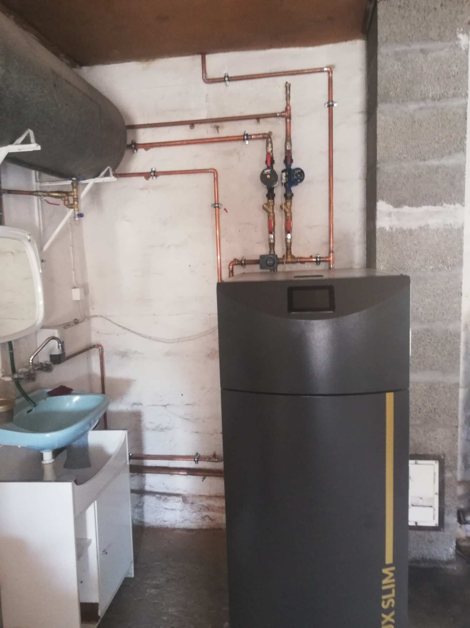 Instalacja kotła grzewczego z miedzianymi rurami w pomieszczeniu gospodarczym. Widoczny zbiornik, umywalka i surowe ściany z betonu komórkowego.