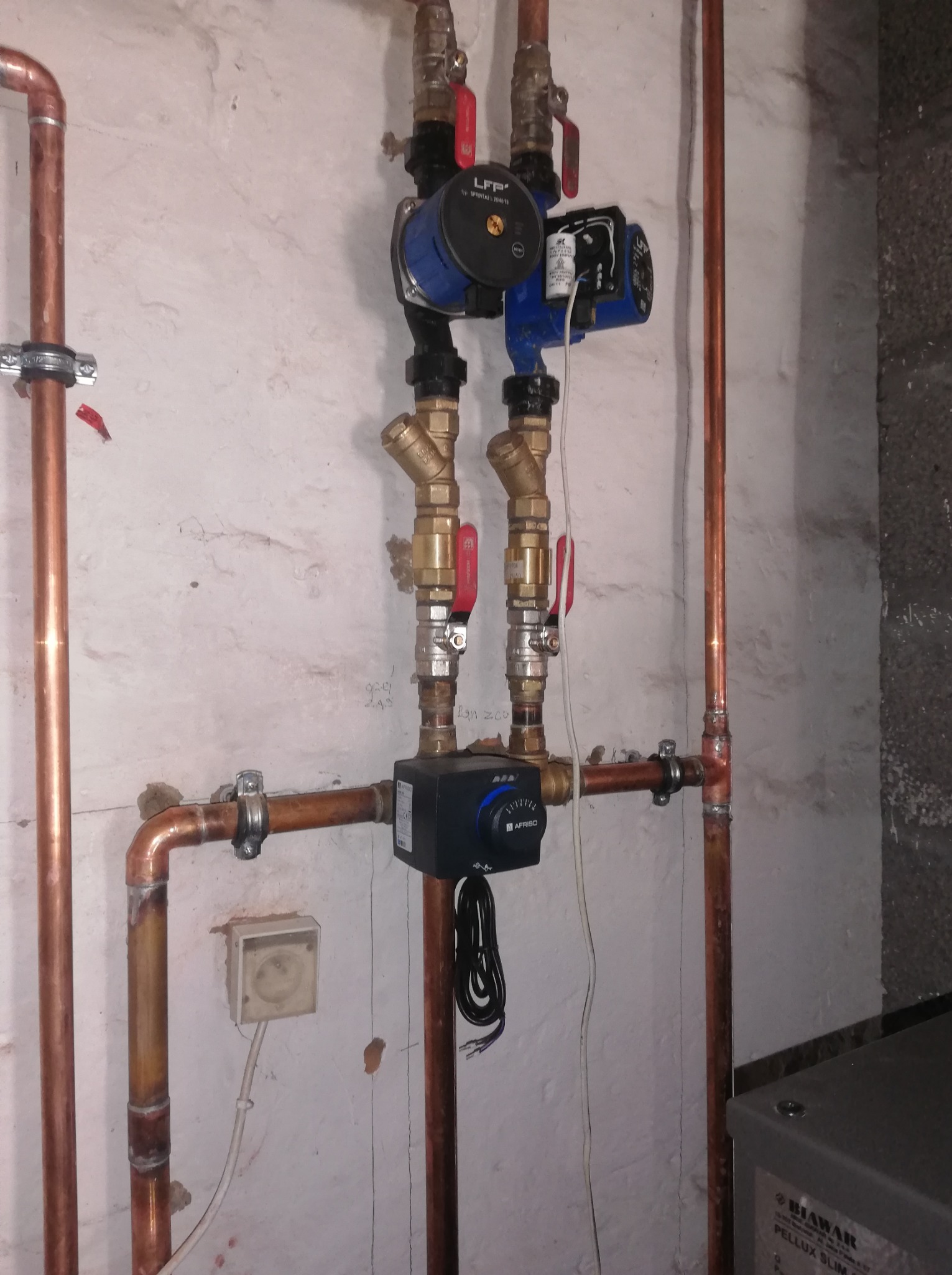 Zmontowana instalacja grzewcza z miedzianymi rurami, zaworami i pompą obiegową LFP na tle ściany z widocznymi niedoskonałościami. Widoczny fragment pieca Biawar.