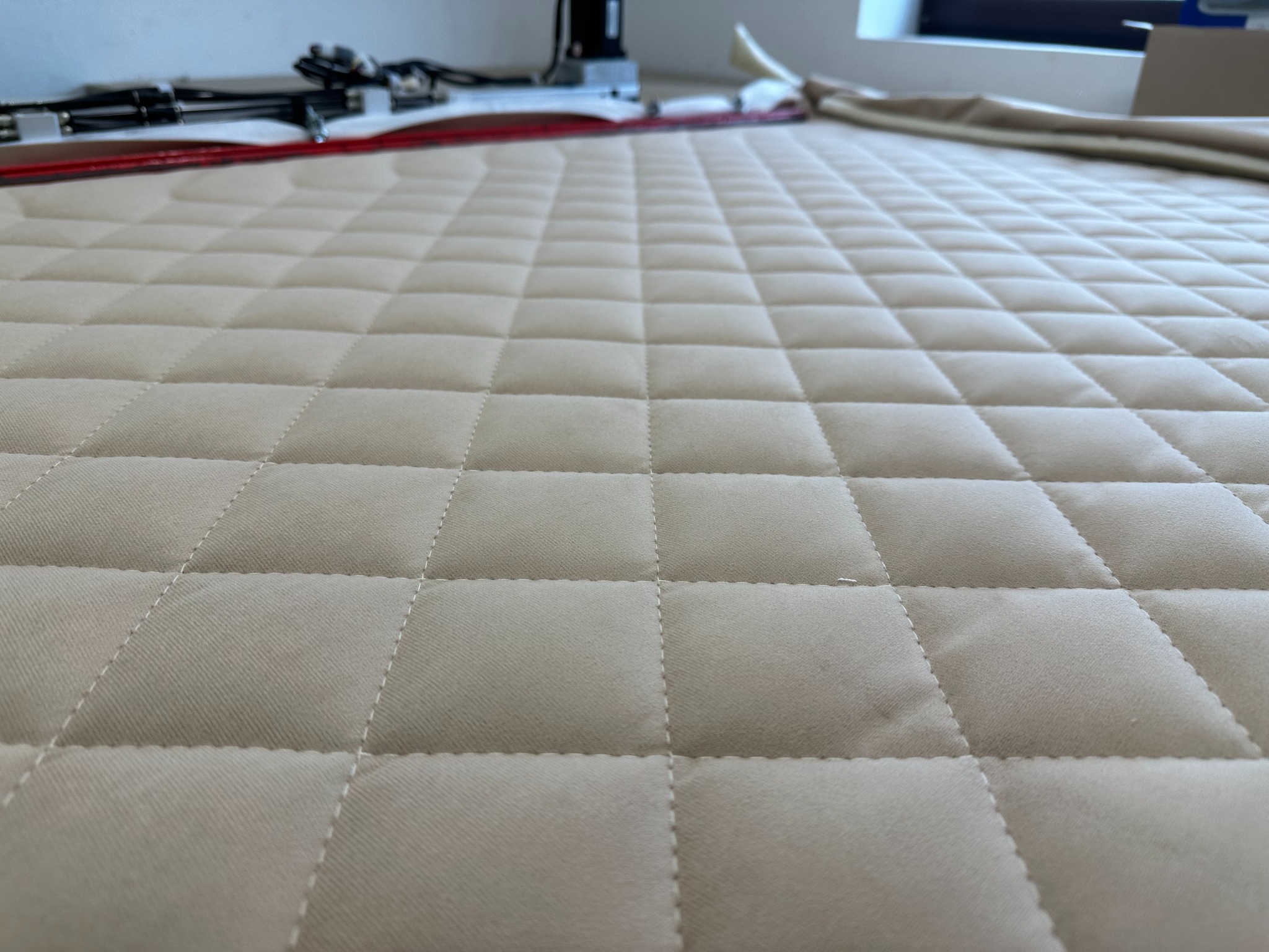 Beżowa, pikowana tkanina z regularnym wzorem rombów, widoczne białe szwy, w tle narzędzia i materiały.