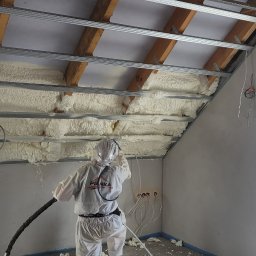 PIANKA PRO IZOLACJE NATRYSKOWE MARCIN GAWEŁKO - Pracownik w kombinezonie ochronnym aplikuje białą pianę izolacyjną na poddaszu z widoczną konstrukcją metalową i drewnianą, otwory elektryczne w ścianie.