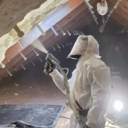 Poddasze w trakcie natrysku pianki przez pracownika w kombinezonie ochronnym. Widoczna drewniana konstrukcja dachu i świeżo nałożona pianka. Oświetlenie halogenowe.