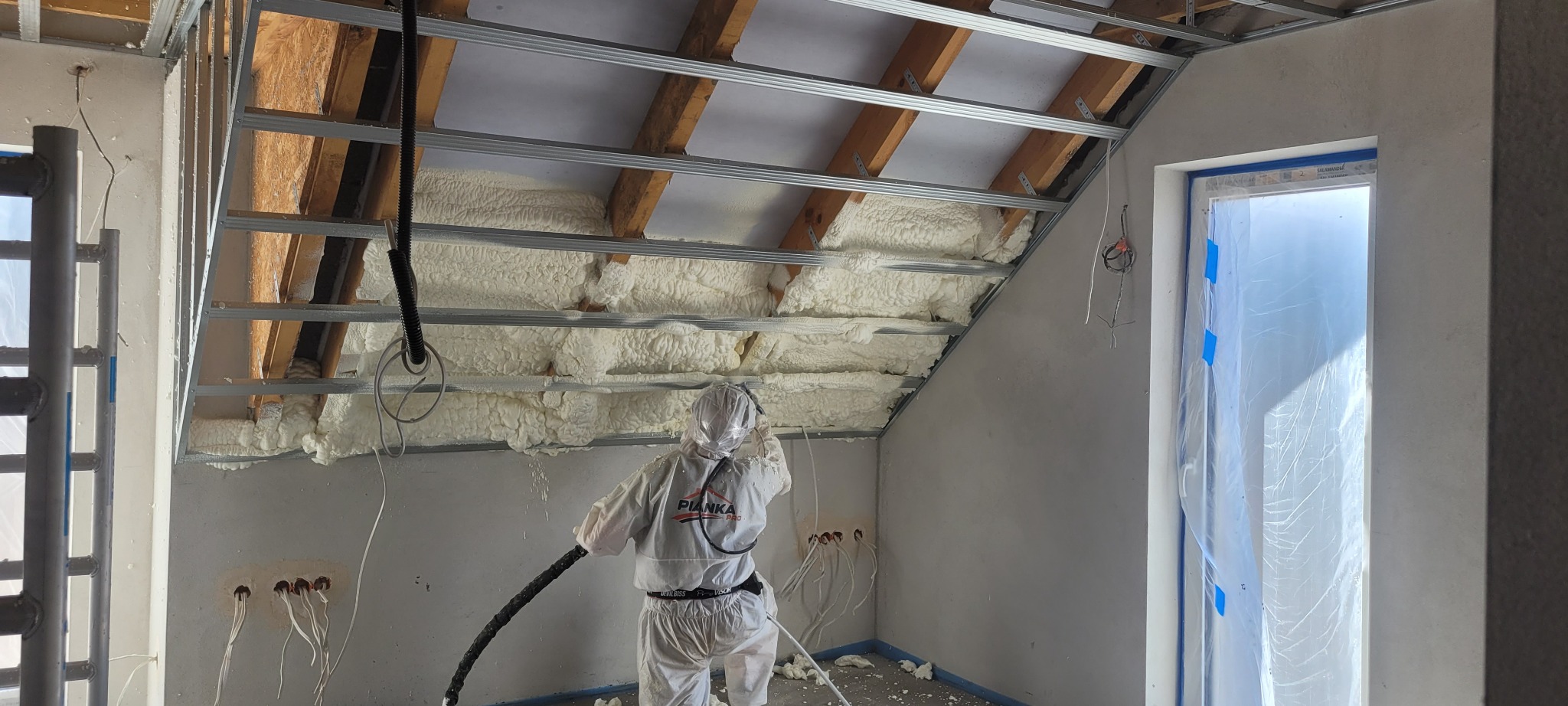 Pracownik w kombinezonie ochronnym aplikuje białą pianę izolacyjną na poddaszu z widoczną konstrukcją metalową i drewnianą, otwory elektryczne w ścianie.