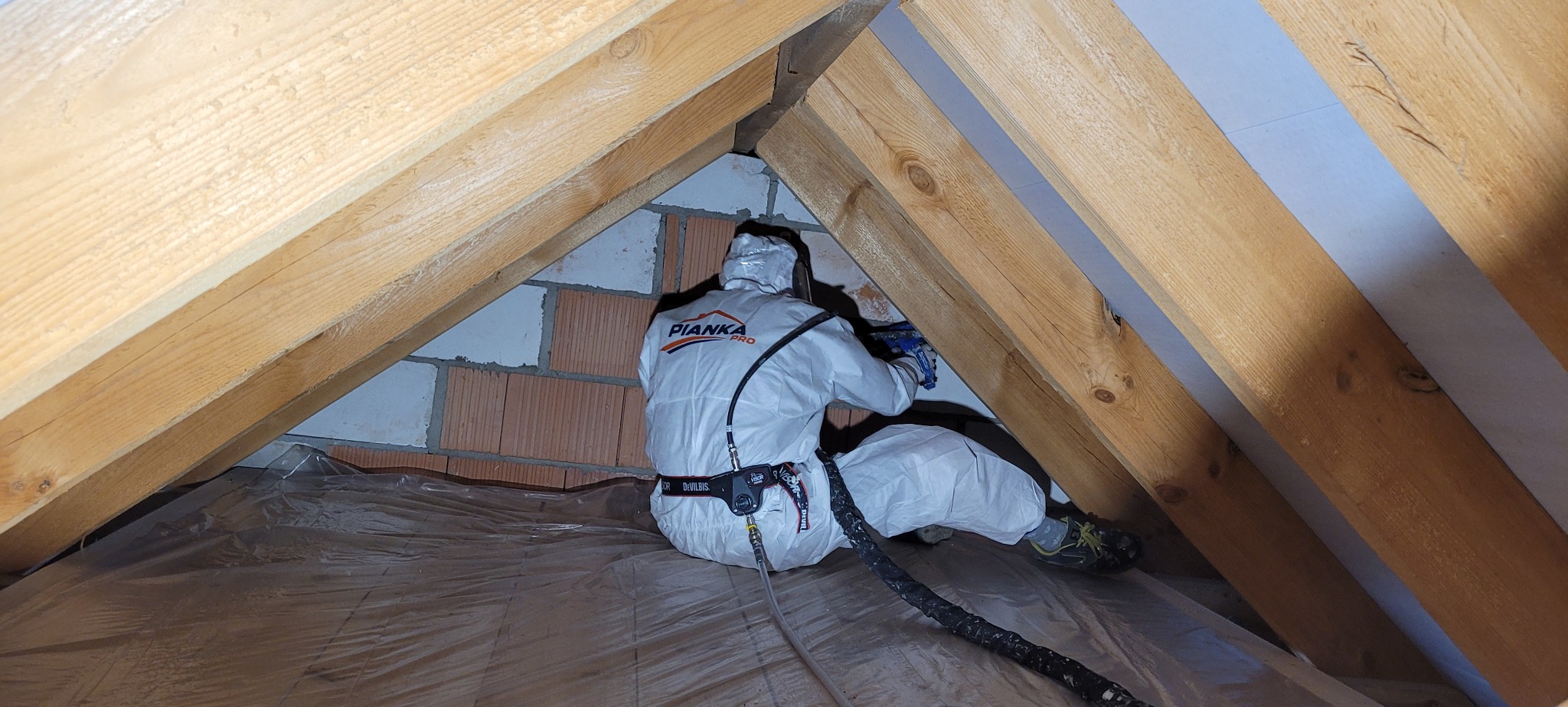 Pracownik w kombinezonie ochronnym z logo 'PIANKA PRO' aplikuje piankę w trudno dostępnym miejscu na poddaszu, widoczne drewniane krokwie i ceglana ściana