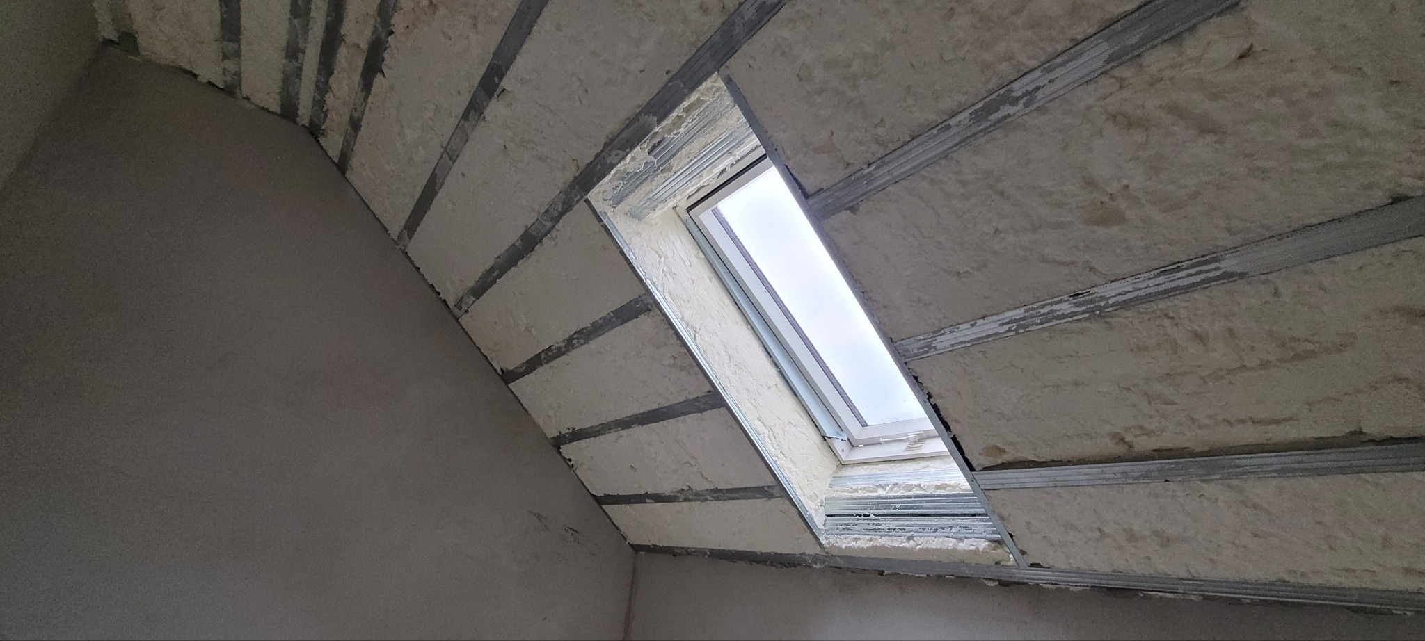 Poddasze z oknem dachowym w trakcie ocieplania: widoczne metalowe profile, wypełnienie jasną pianką izolacyjną oraz fragment nieocieplonej ściany.