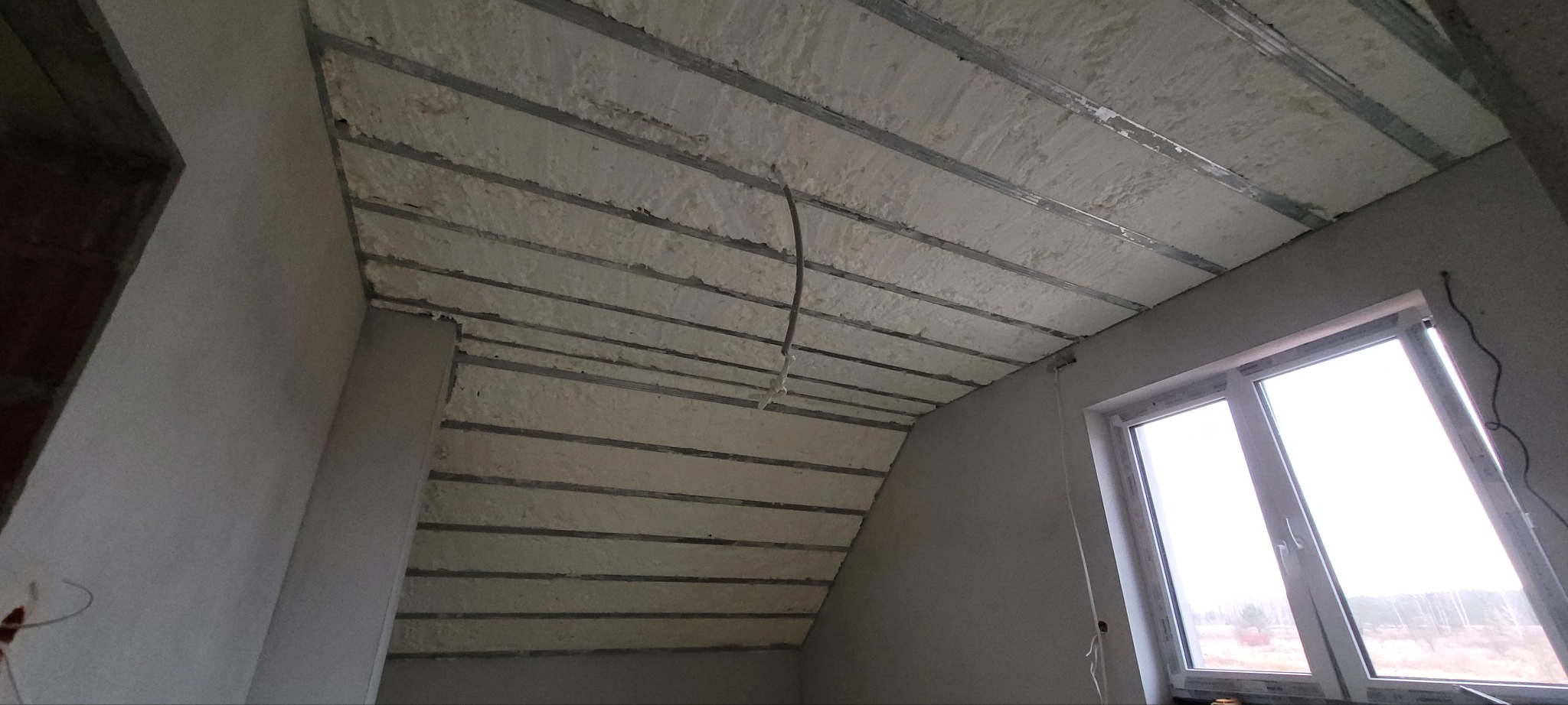 Poddasze z widoczną konstrukcją stalową i nałożoną warstwą izolacji natryskowej, okno w ścianie szczytowej, instalacja elektryczna w trakcie montażu.