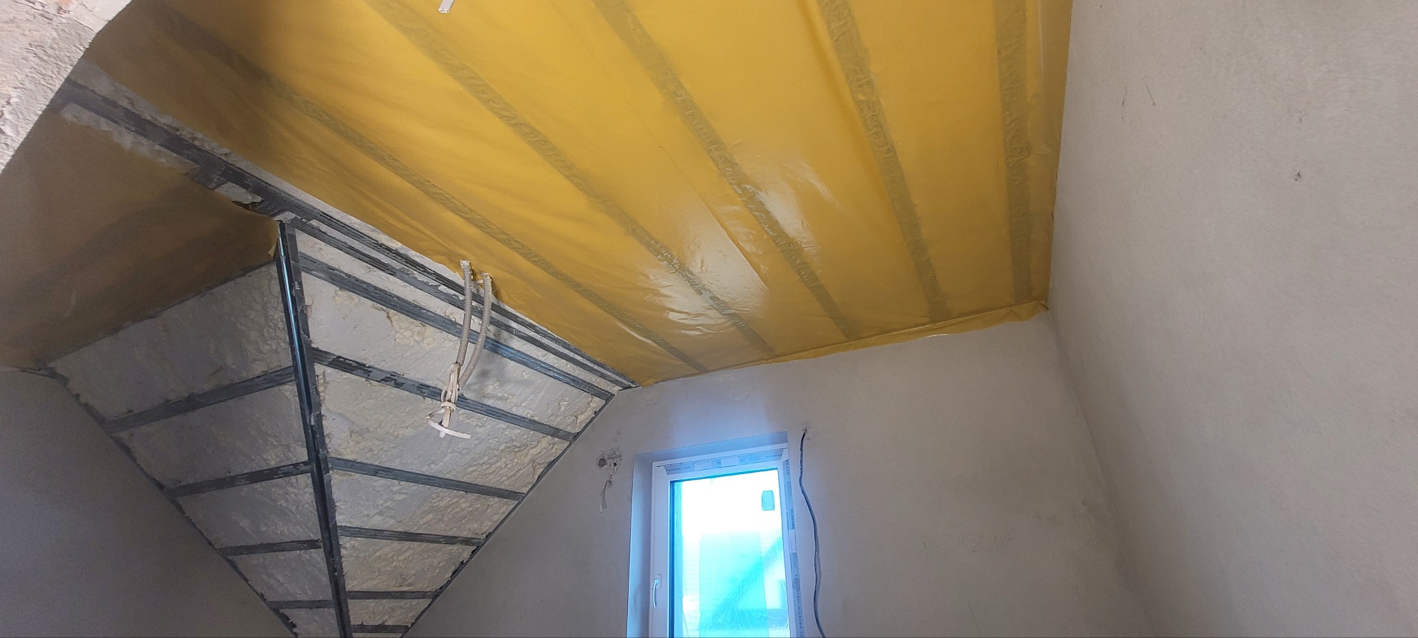 Poddasze w trakcie ocieplania: widoczne metalowe profile, wypełnienie izolacyjne i żółta folia paroizolacyjna, okno w ścianie bocznej.