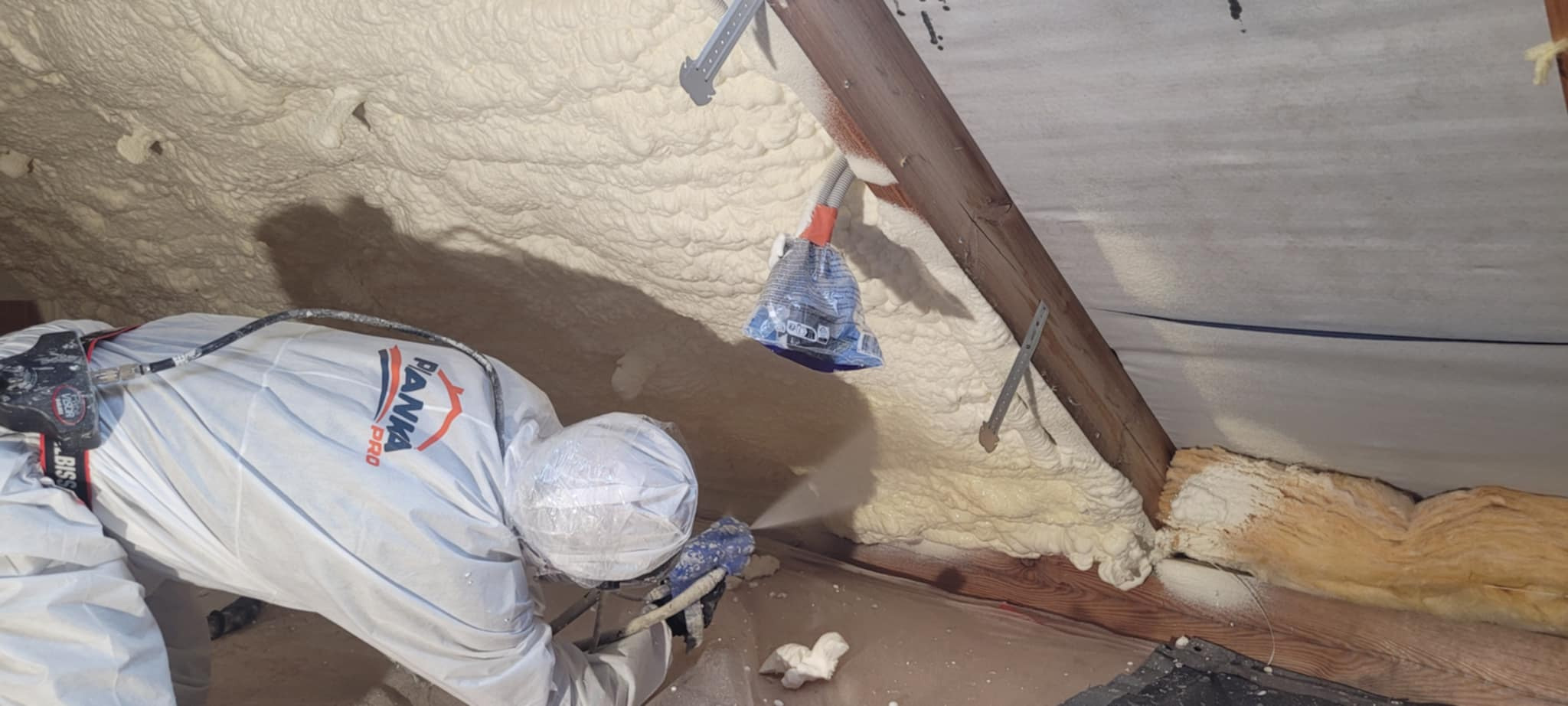 Ocieplanie pianką PUR poddasza w Białymstoku. Pracownik w kombinezonie aplikuje pianę na skośną powierzchnię, widoczna struktura izolacji i elementy konstrukcji dachu.