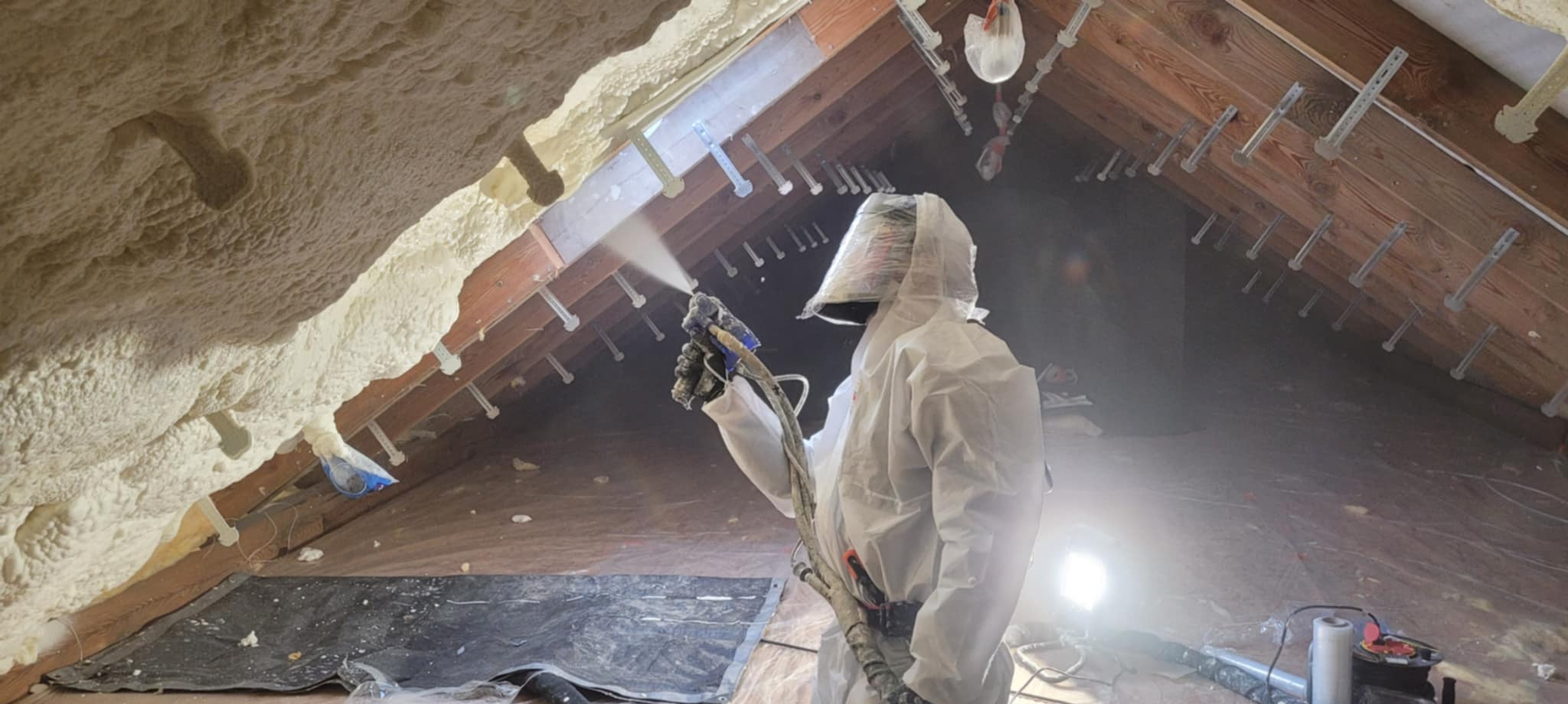 Poddasze w trakcie natrysku pianki przez pracownika w kombinezonie ochronnym. Widoczna drewniana konstrukcja dachu i świeżo nałożona pianka. Oświetlenie halogenowe.