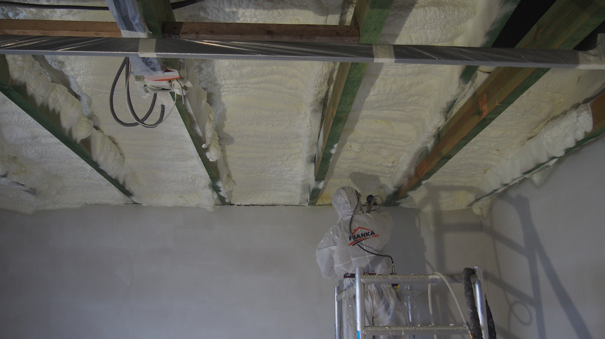 Aplikacja białej pianki izolacyjnej na poddaszu przez pracownika w kombinezonie ochronnym. Widoczne drewniane belki i instalacje.
