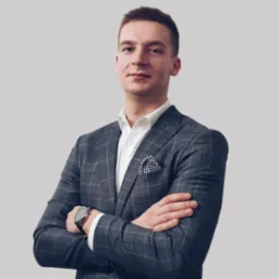 Młody mężczyzna w szarej, kraciastej marynarce i białej koszuli, z zegarkiem na ręku i poszetką w kieszonce, pozuje na jasnym tle z założonymi rękami.