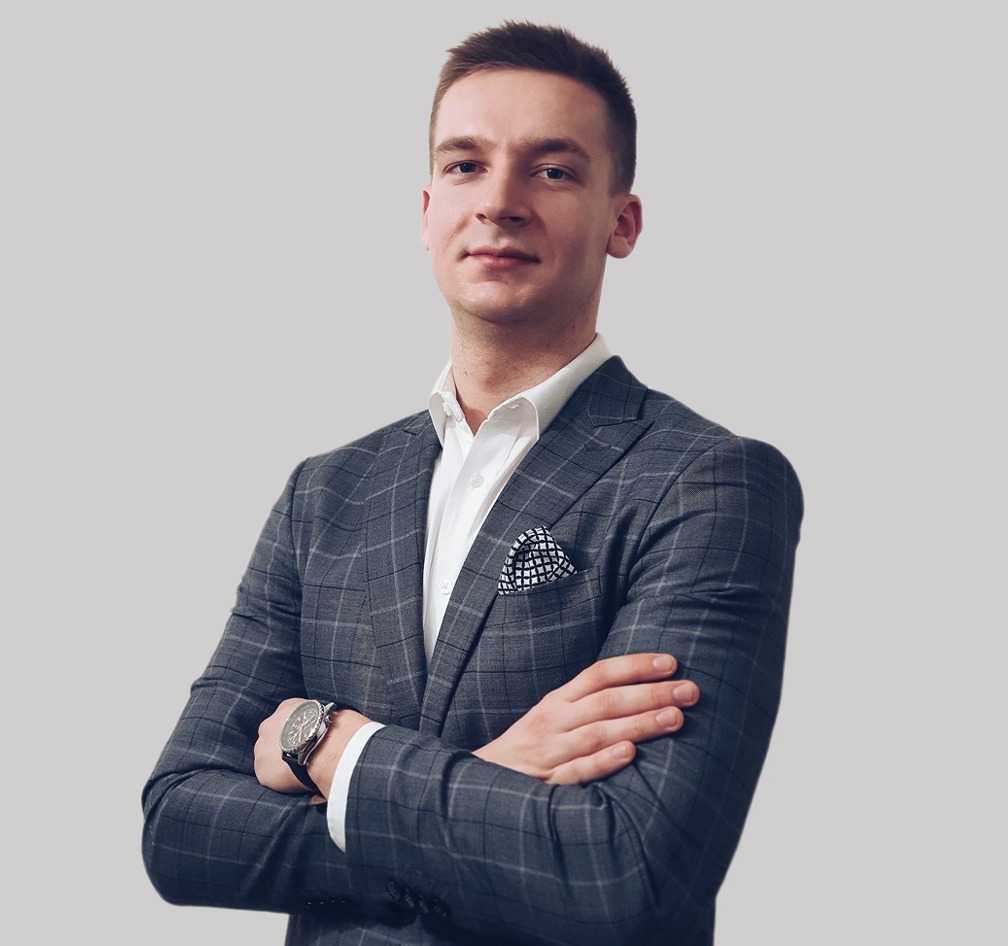 Młody mężczyzna w szarej, kraciastej marynarce i białej koszuli, z zegarkiem na ręku i poszetką w kieszonce, pozuje na jasnym tle z założonymi rękami.