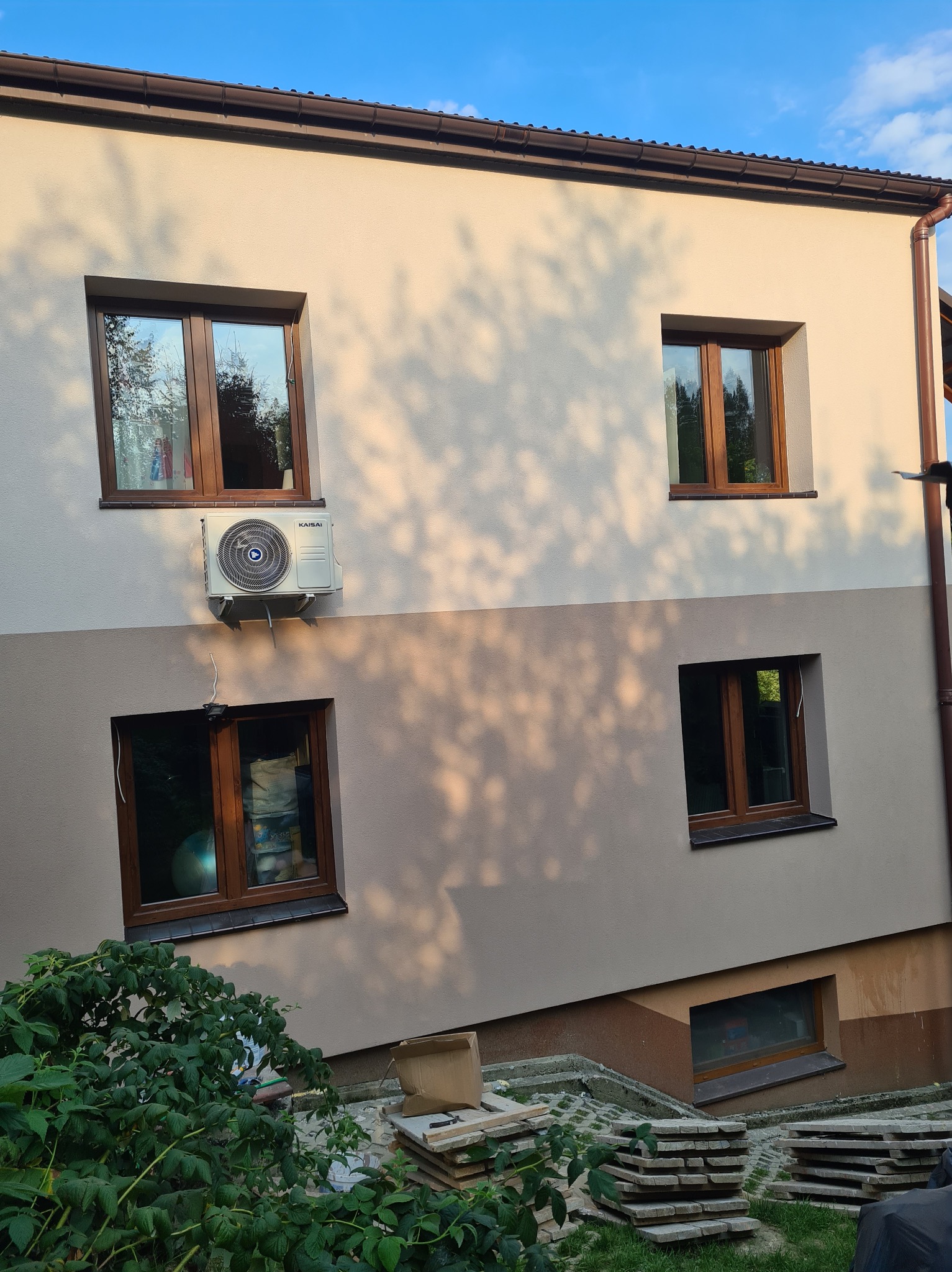 Dwukolorowa elewacja domu z brązowymi oknami, klimatyzatorem Kaisai i stertą płyt chodnikowych przy krzewach.