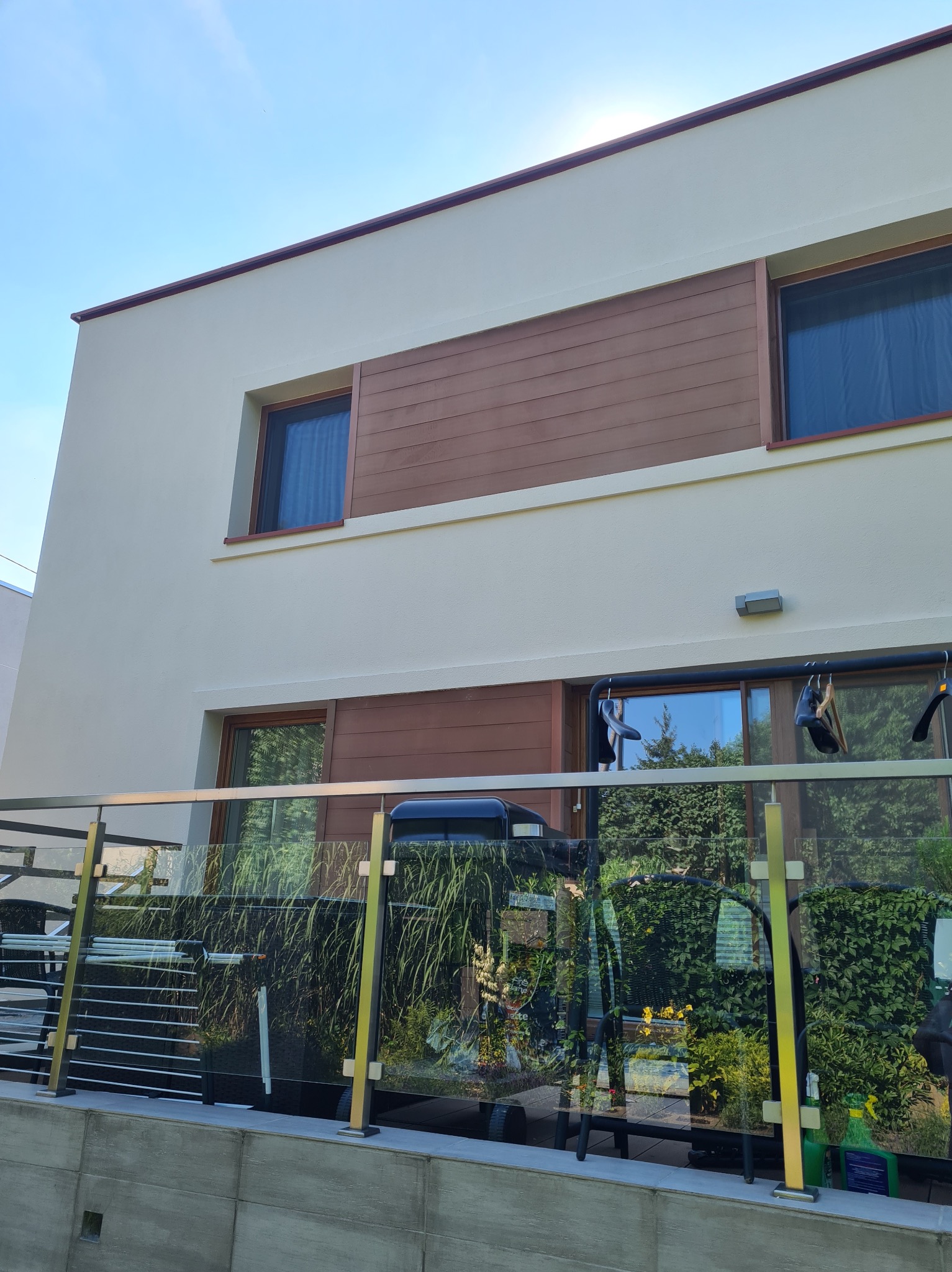 Dwukondygnacyjny dom z kremową elewacją i poziomymi panelami w kolorze drewna, widoczny z poziomu tarasu z szklaną balustradą, na której wiszą wieszaki na ubrania.