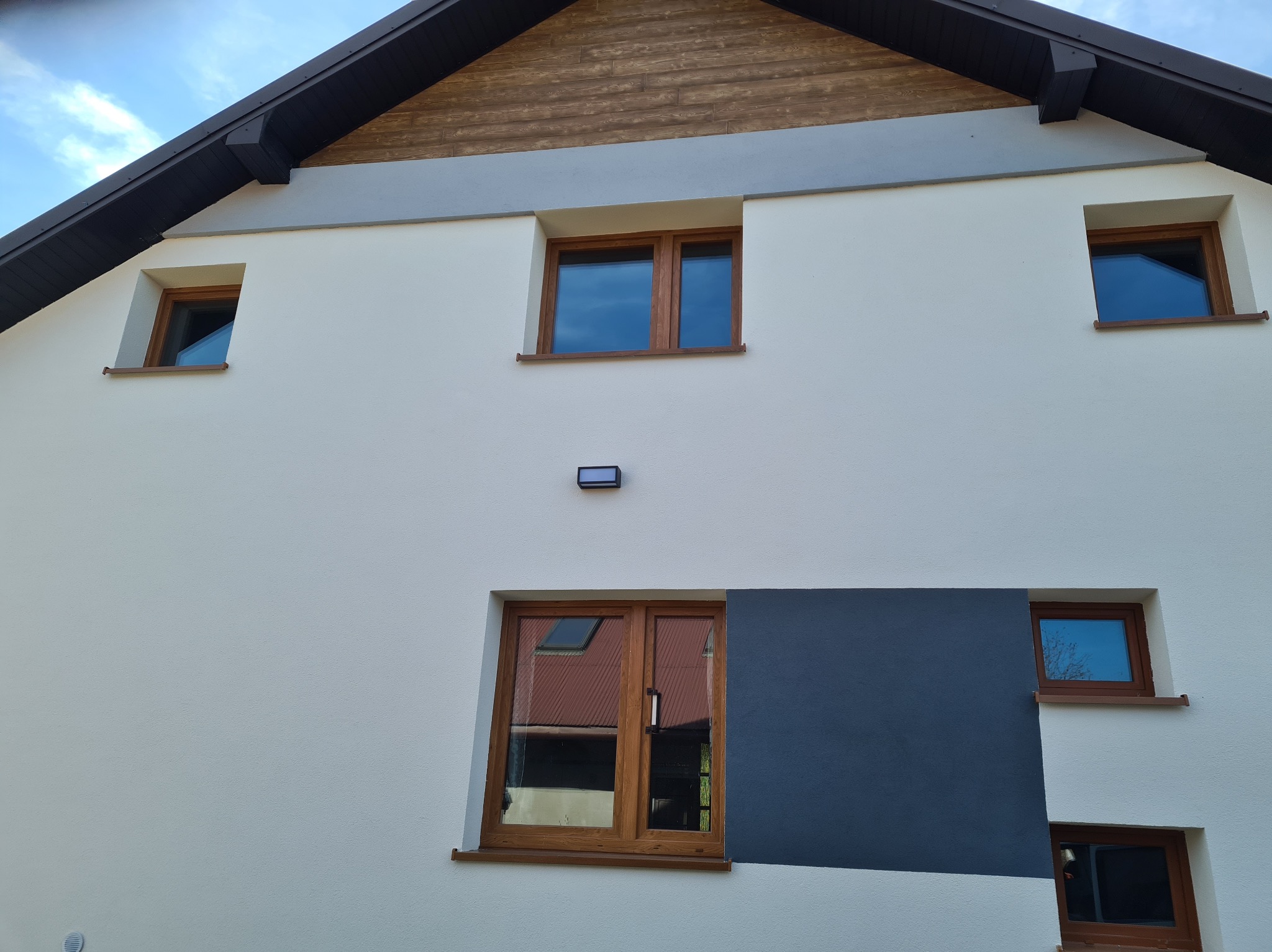 Nowoczesna elewacja domu jednorodzinnego z drewnianymi oknami, fragment ściany pomalowany na grafitowo, widoczny fragment dachu i nieba.